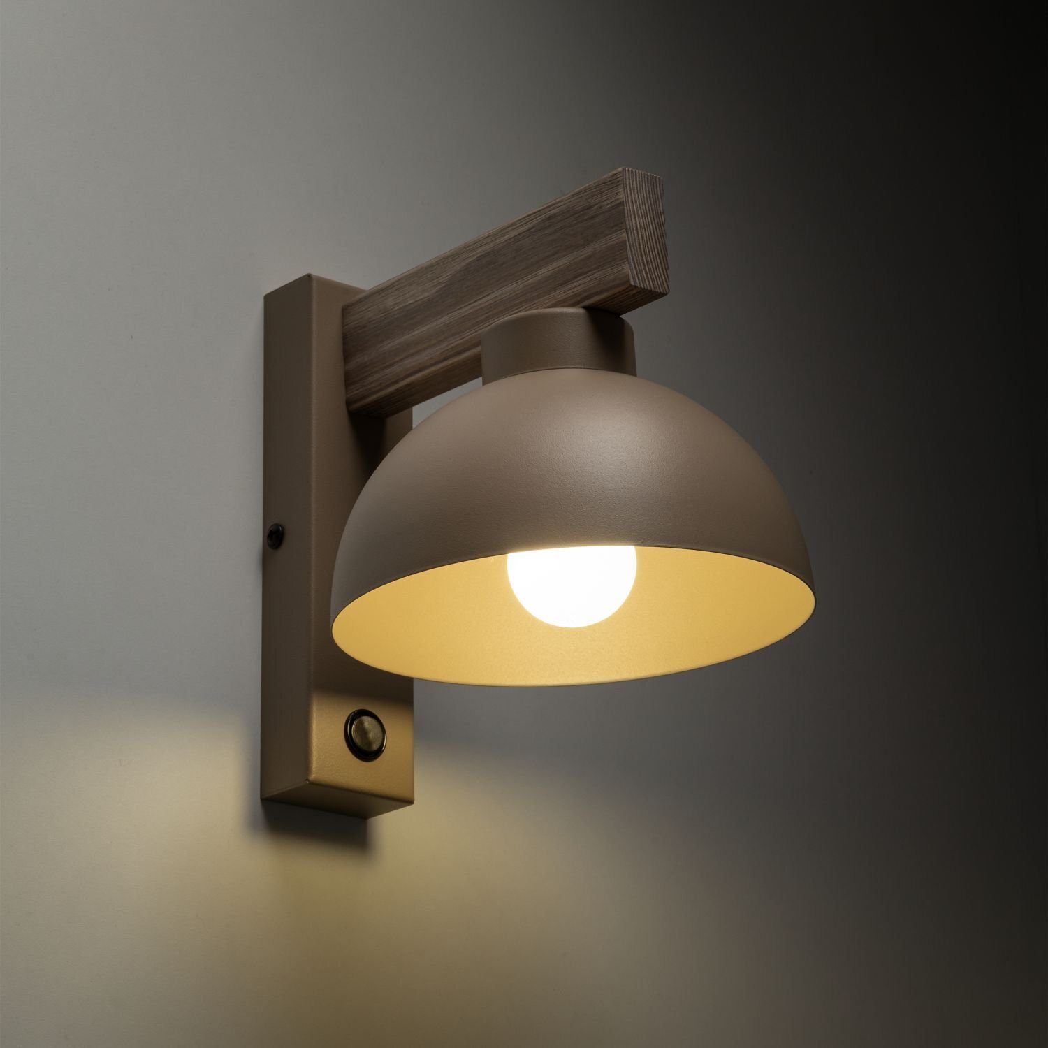 Beżowy kinkiet ścienny z włącznikiem, posiadający półokrągły klosz i drewniany element mocujący. Lampa jest włączona, emitując ciepłe światło.