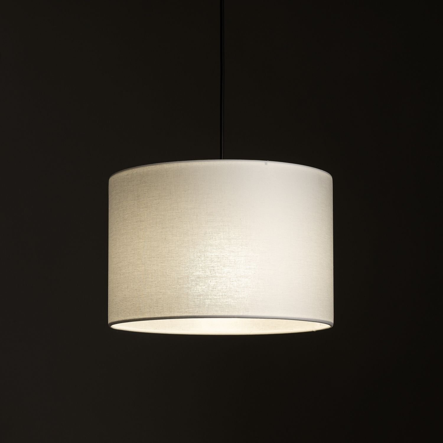 Na obrazie widoczna jest biała, cylindryczna lampa wisząca z materiałowym abażurem, emitująca ciepłe światło. Tło jest ciemne, co podkreśla jasność lampy.