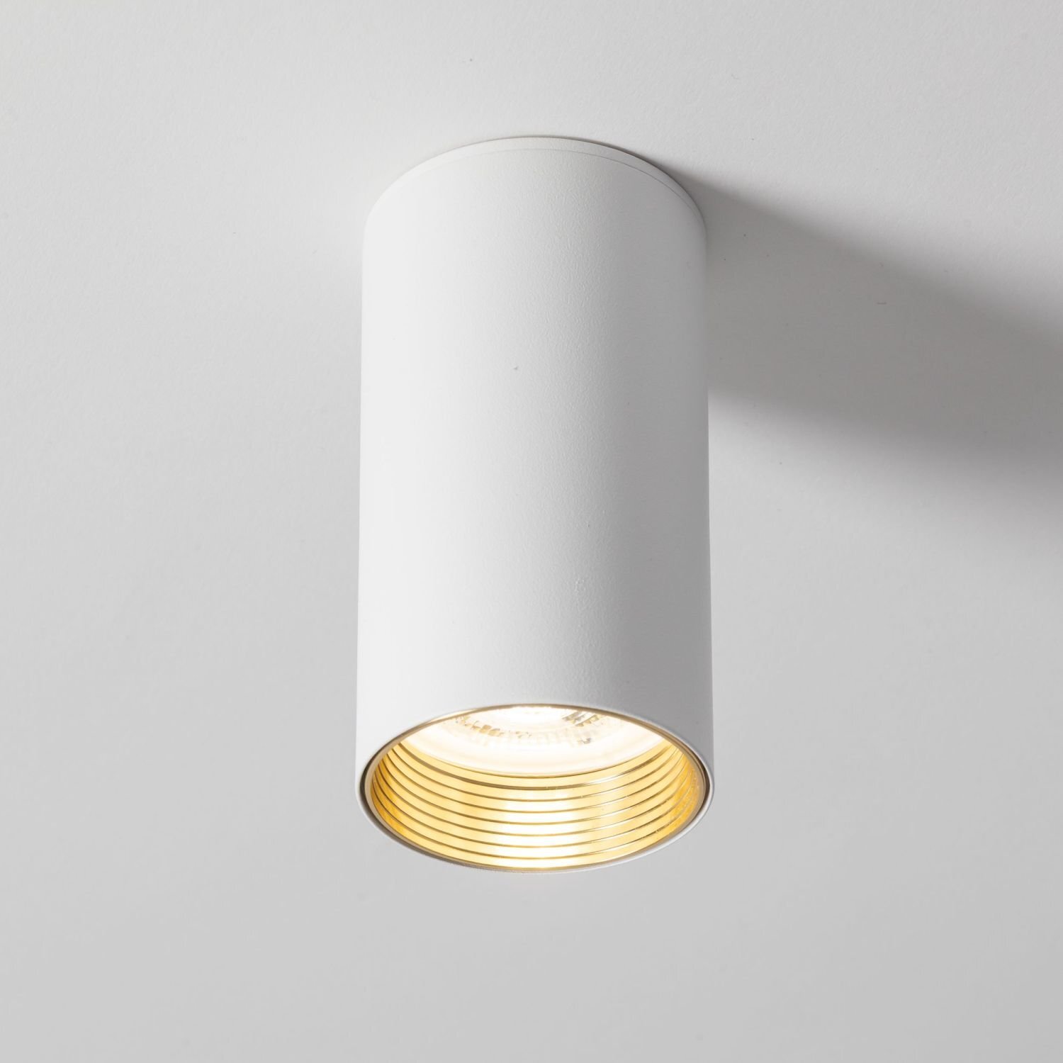Biały, cylindryczny spot natynkowy z widocznym złotym wnętrzem. Lampa jest zamontowana na suficie.