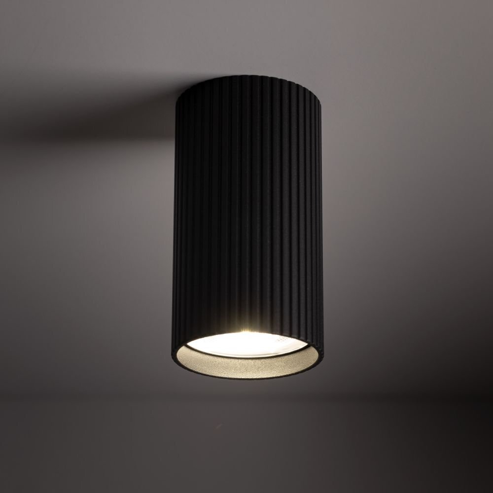 Czarna, cylindryczna oprawa natynkowa z żebrowaną powierzchnią, emitująca światło od dołu. Zamontowana na suficie w nowoczesnym wnętrzu.