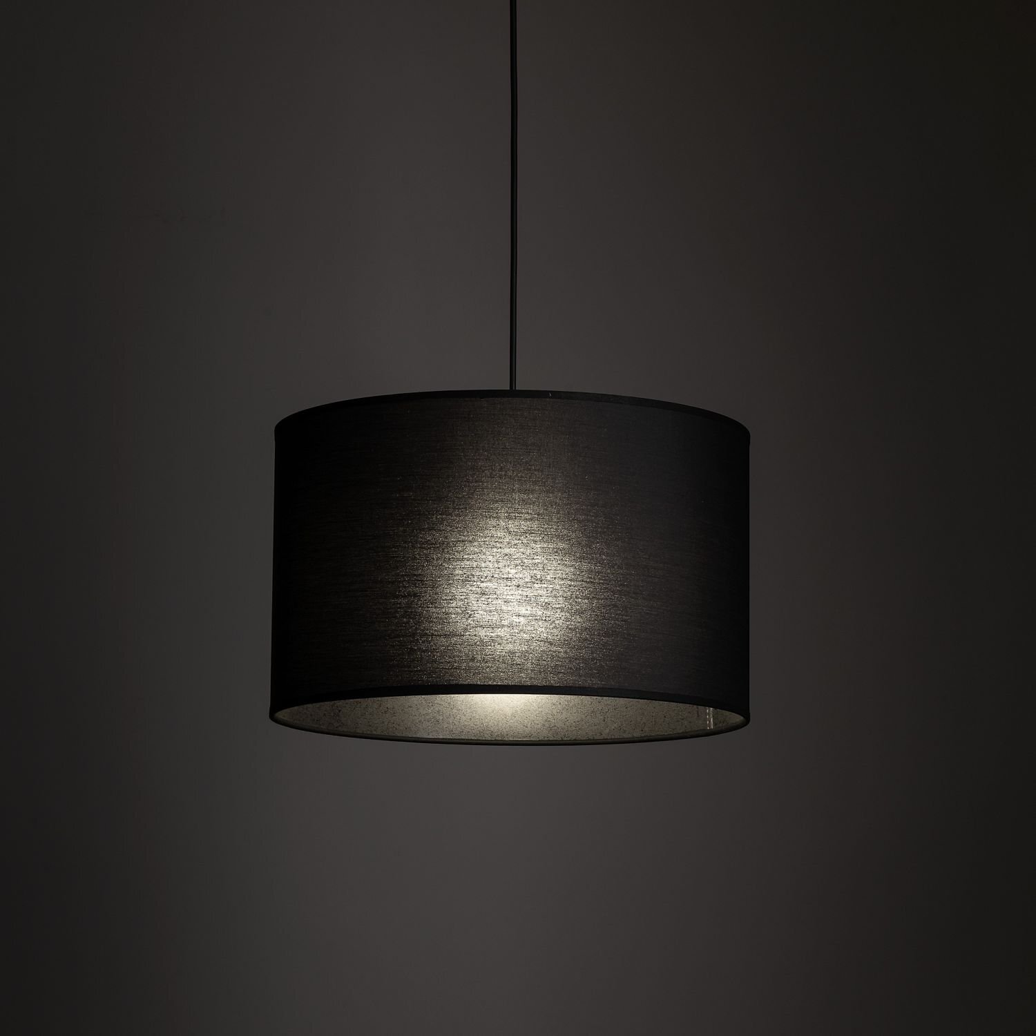 Czarna lampa wisząca z cylindrycznym abażurem, emitująca delikatne światło. Lampa zawieszona jest na cienkim przewodzie na tle ciemnego otoczenia.