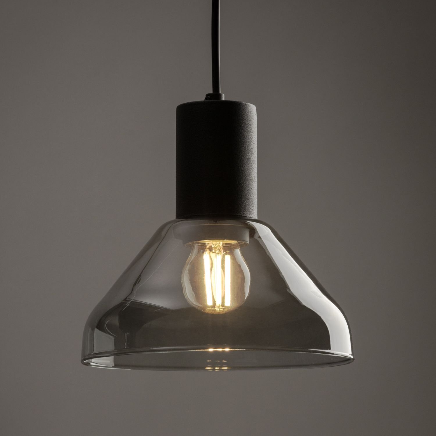 Lampa wisząca z czarnym, cylindrycznym uchwytem i przezroczystym kloszem, wewnątrz którego znajduje się żarówka. Światło żarówki jest ciepłe i delikatnie oświetla wnętrze klosza.