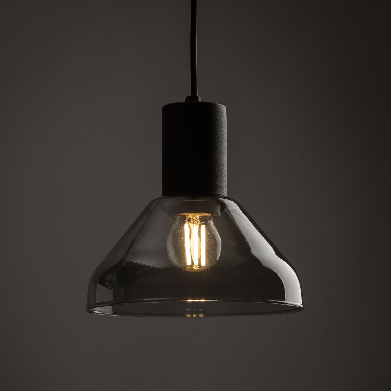 Czarna lampa wisząca z przezroczystym kloszem, wewnątrz której znajduje się żarówka emitująca ciepłe światło. Lampa zawieszona jest na czarnym przewodzie.