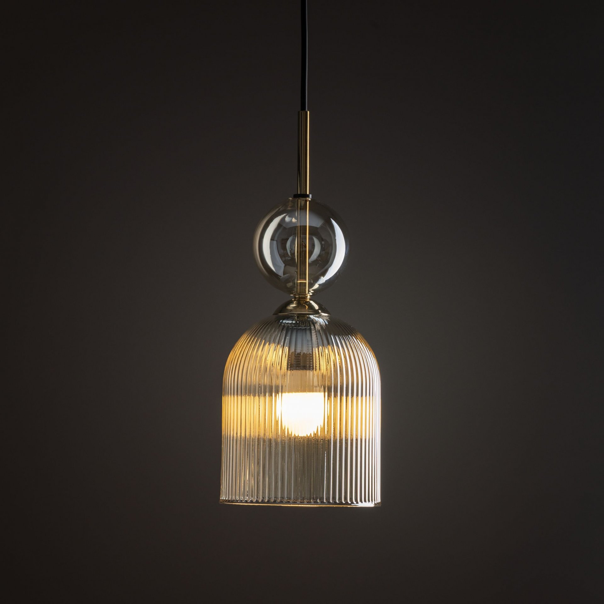 Lampa wisząca z czarną oprawą i bursztynowym kloszem, emitująca ciepłe światło. Klosz ma żłobioną powierzchnię, a nad nim znajduje się przezroczysta kula.