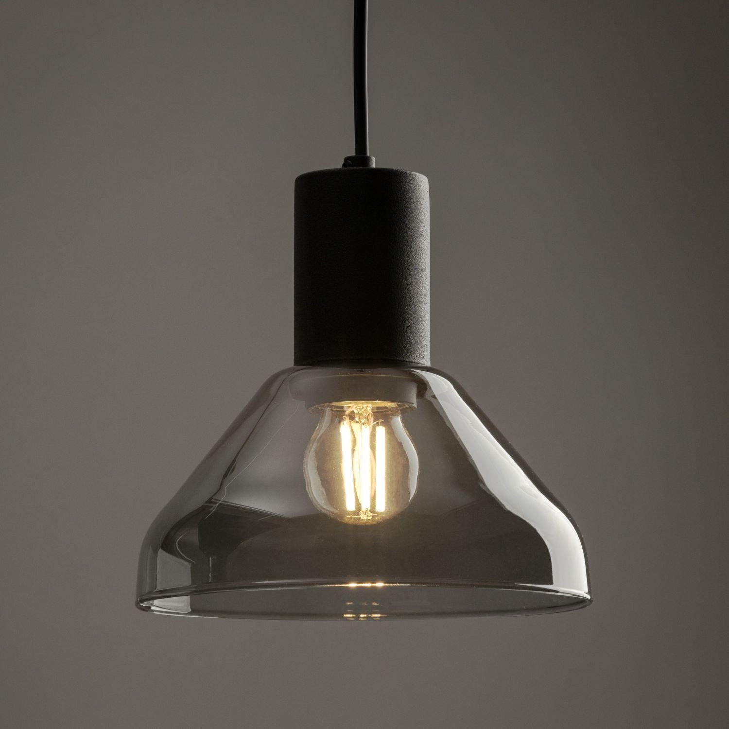 Czarna lampa wisząca z przezroczystym kloszem, w której widoczna jest żarówka emitująca ciepłe światło. Lampa ma prosty, nowoczesny design.