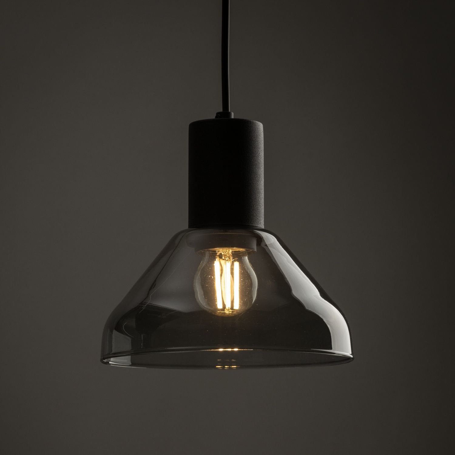 Czarna lampa wisząca z przezroczystym kloszem, wewnątrz której znajduje się żarówka emitująca ciepłe światło. Lampa zawieszona jest na czarnym przewodzie.
