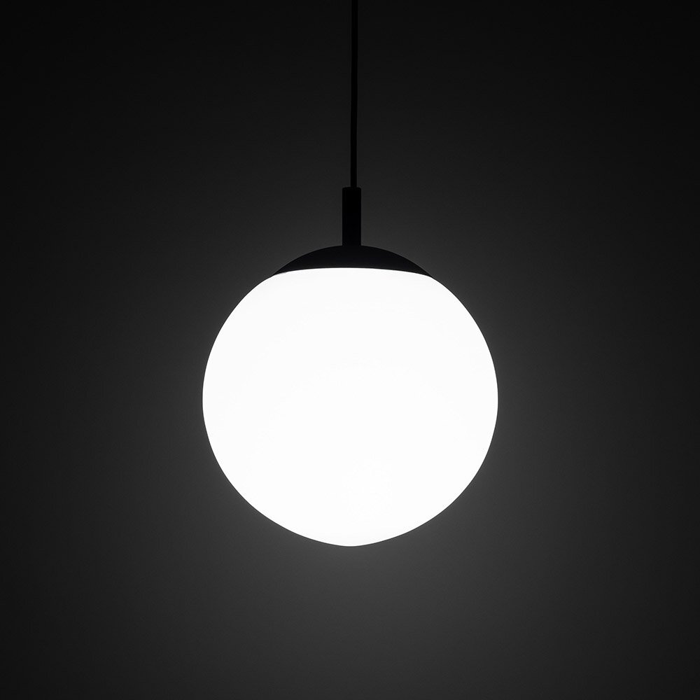 Na obrazie widoczna jest wisząca lampa o kulistym, białym kloszu, zawieszona na cienkim przewodzie. Tło jest ciemne, co podkreśla jasność lampy.