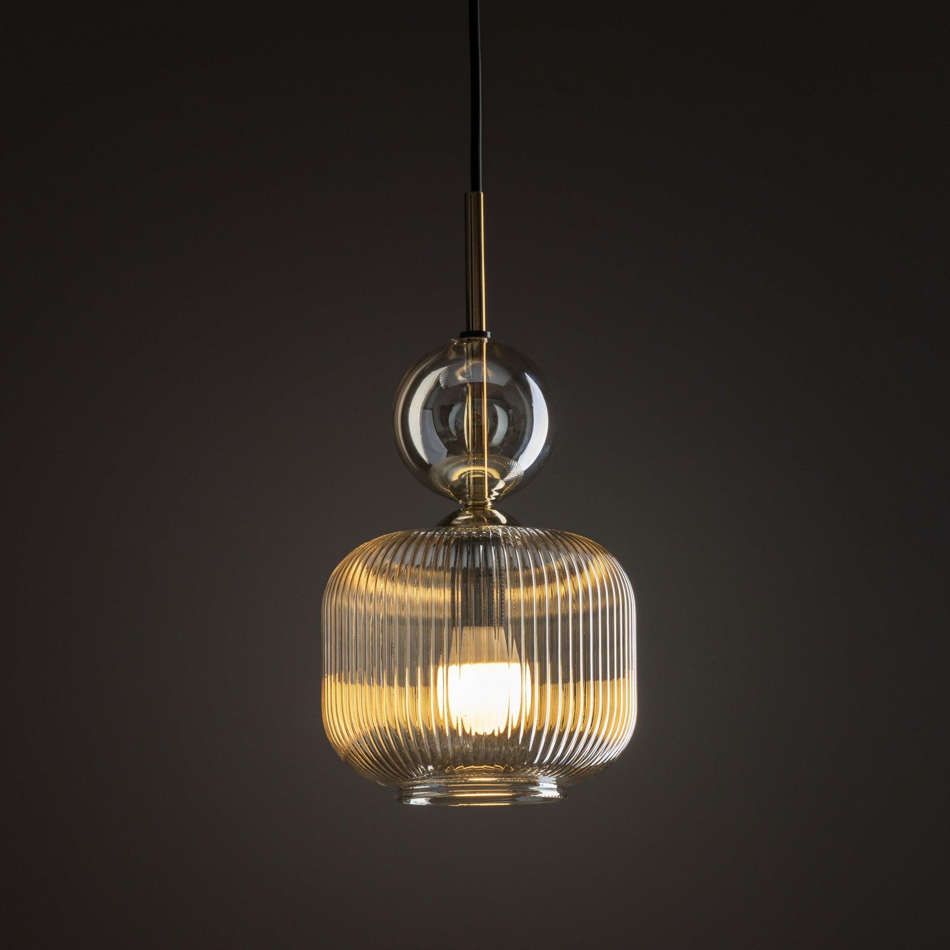 Lampa wisząca z bursztynowym kloszem o żebrowanej strukturze, zawieszona na czarnym przewodzie. Światło emituje ciepły blask, a nad kloszem znajduje się przezroczysta kula.