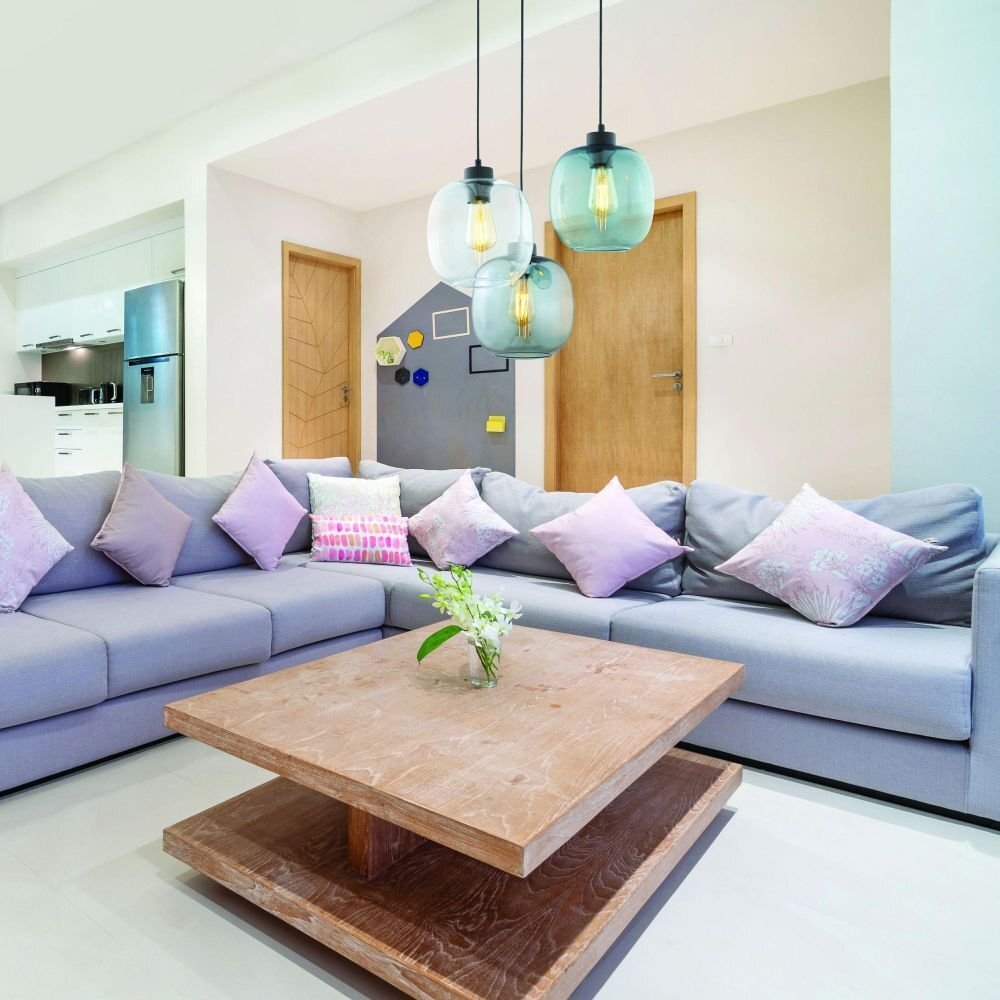 W salonie znajduje się szara sofa z fioletowymi poduszkami oraz drewniany stolik kawowy. Nad stolikiem wiszą trzy nowoczesne lampy z przezroczystymi kloszami w kolorze niebieskim.