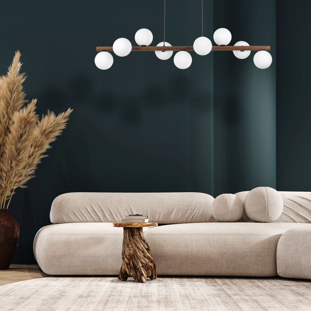 Na obrazie widoczna jest nowoczesna lampa wisząca z dziewięcioma kulistymi kloszami, zawieszona nad jasną sofą. W tle znajduje się ciemna ściana oraz dekoracyjna roślina w dużej donicy.