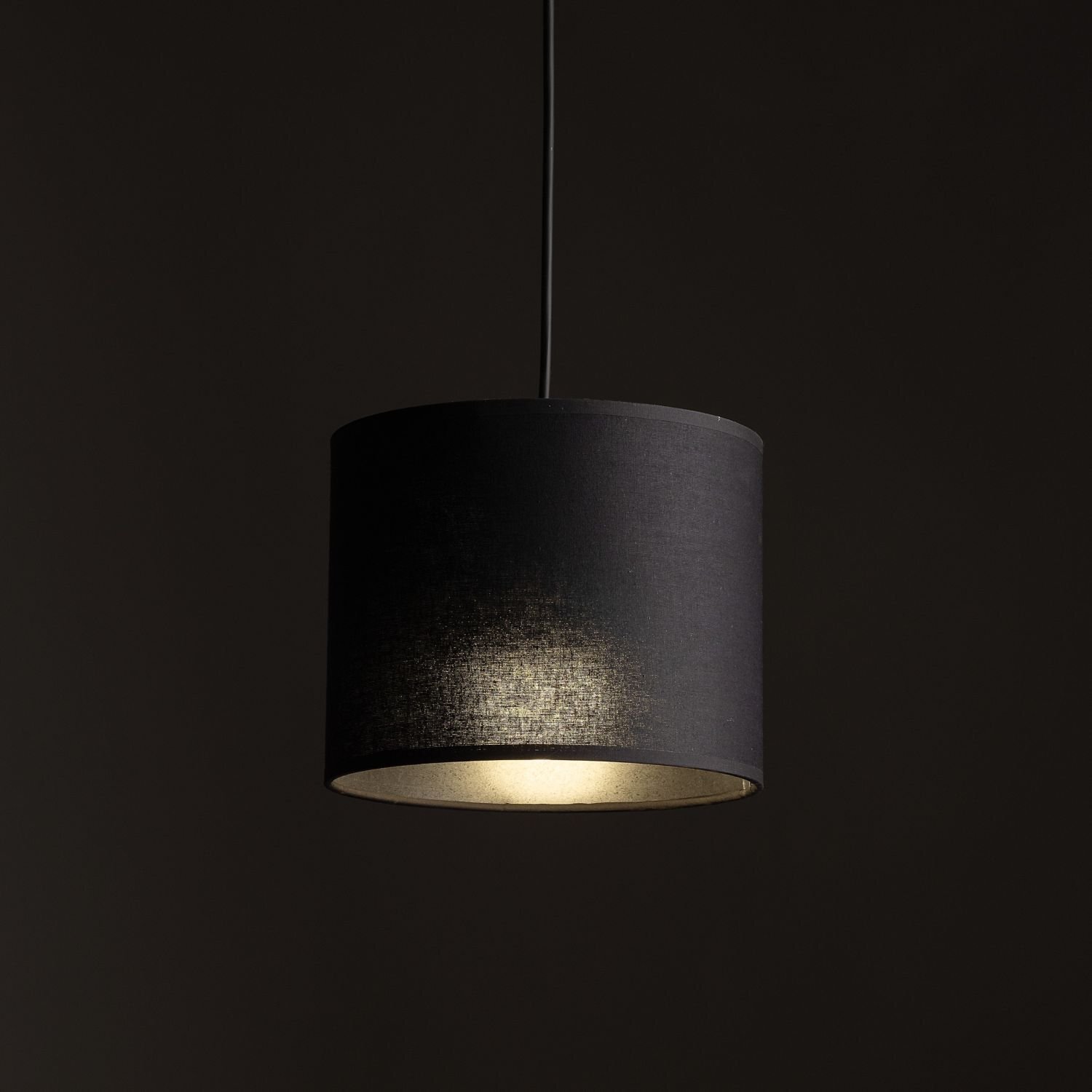 Czarna, cylindryczna lampa wisząca z materiałowym abażurem, emitująca ciepłe światło. Lampa zawieszona jest na cienkim przewodzie na ciemnym tle.