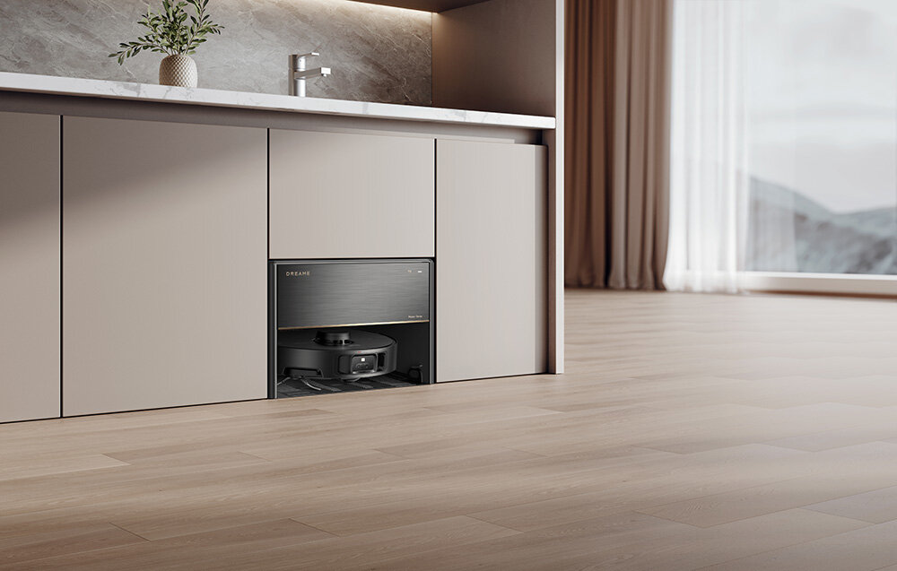 Wnętrze nowoczesnej kuchni z szafkami, w jednej z nich znajduje się robot sprzątający. Na blacie stoi wazon z rośliną, a w tle widoczne jest duże okno z zasłonami.