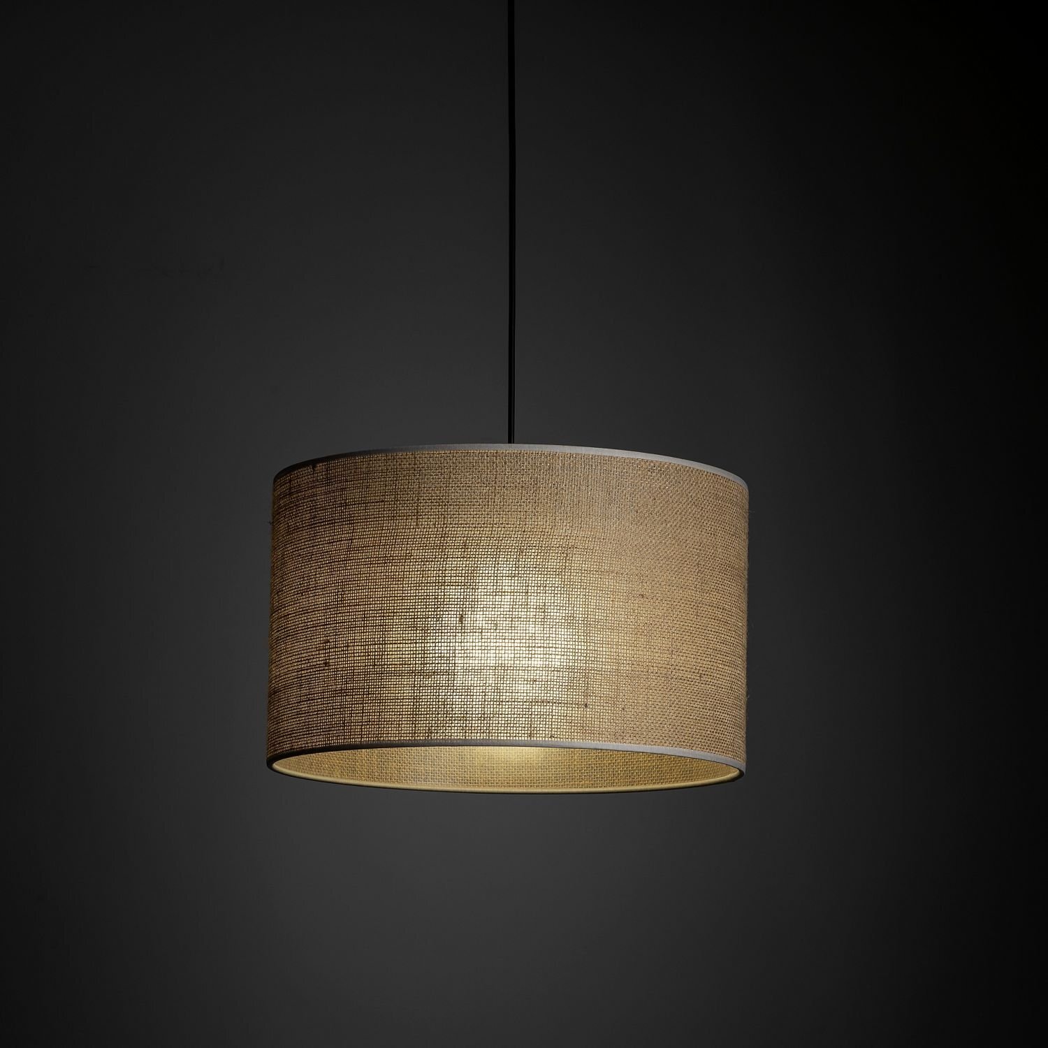 Na obrazie widoczna jest lampa wisząca z cylindrycznym abażurem w kolorze beżowym, zawieszona na czarnym przewodzie. Tło jest ciemne, co podkreśla oświetlenie lampy.