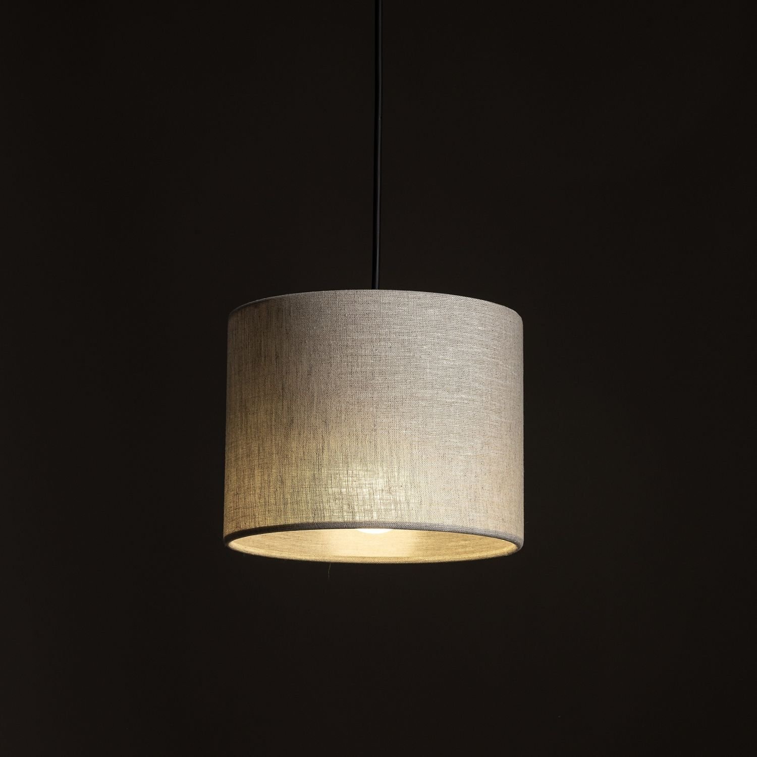 Na obrazie widoczna jest lampa wisząca z cylindrycznym kloszem w kolorze beżowym, wykonanym z lnu. Lampa jest zawieszona na czarnym przewodzie, a tło jest ciemne.