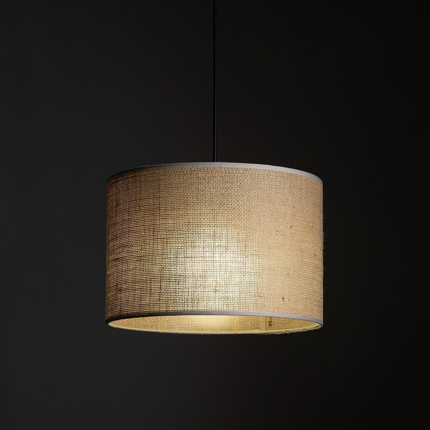 Lampa wisząca z cylindrycznym abażurem w kolorze beżowym, wykonanym z materiału przypominającego jutę. Lampa jest zawieszona na czarnym przewodzie, a tło jest ciemne.
