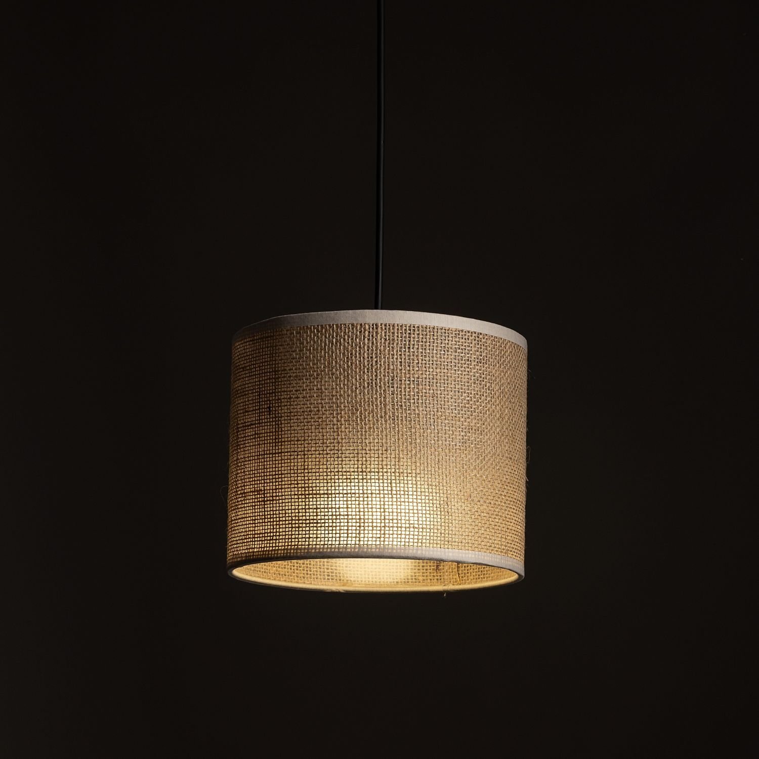 Na ciemnym tle wisi lampa z cylindrycznym abażurem w kolorze beżowym, wykonanym z materiału o teksturze przypominającej tkaninę. Lampa emituje ciepłe, rozproszone światło.