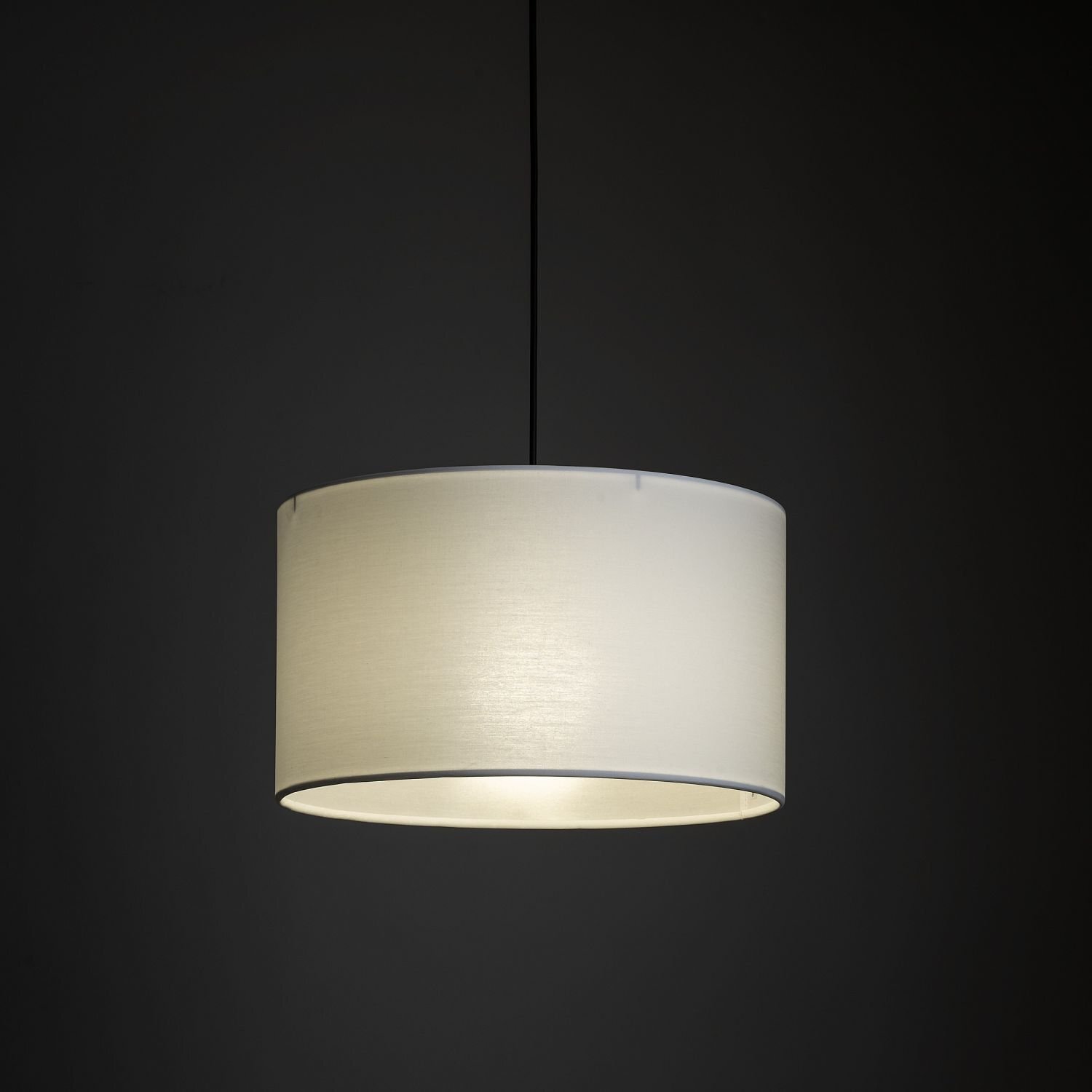 Na ciemnym tle wisi biała, cylindryczna lampa z materiałowym abażurem. Lampa jest podświetlona od wewnątrz, co tworzy delikatne światło.