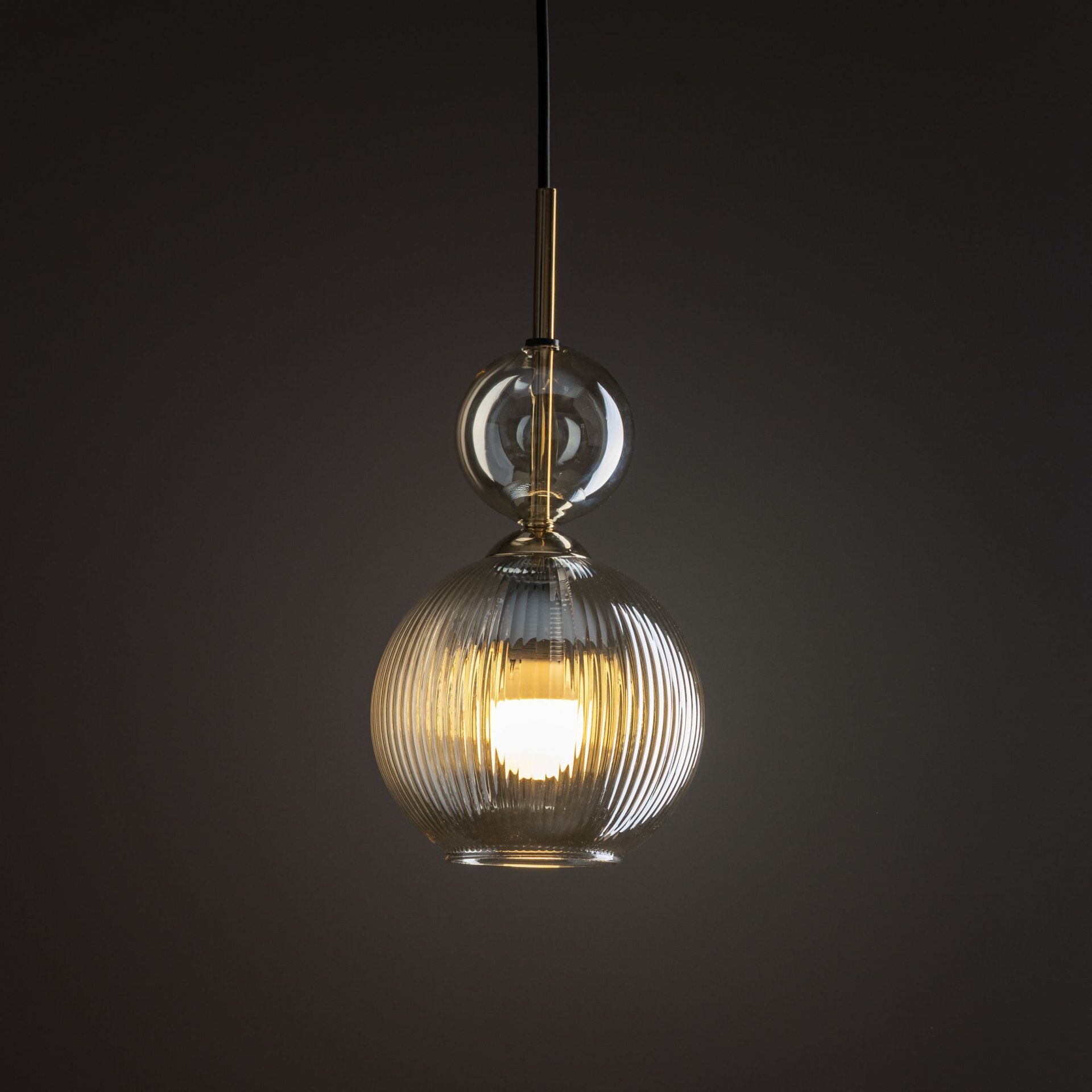 Lampa wisząca z czarnym przewodem i szklanym kloszem w kolorze koniakowym, z widocznym źródłem światła wewnątrz. Klosz ma żłobioną powierzchnię, a nad nim znajduje się mniejsza, przezroczysta kula.