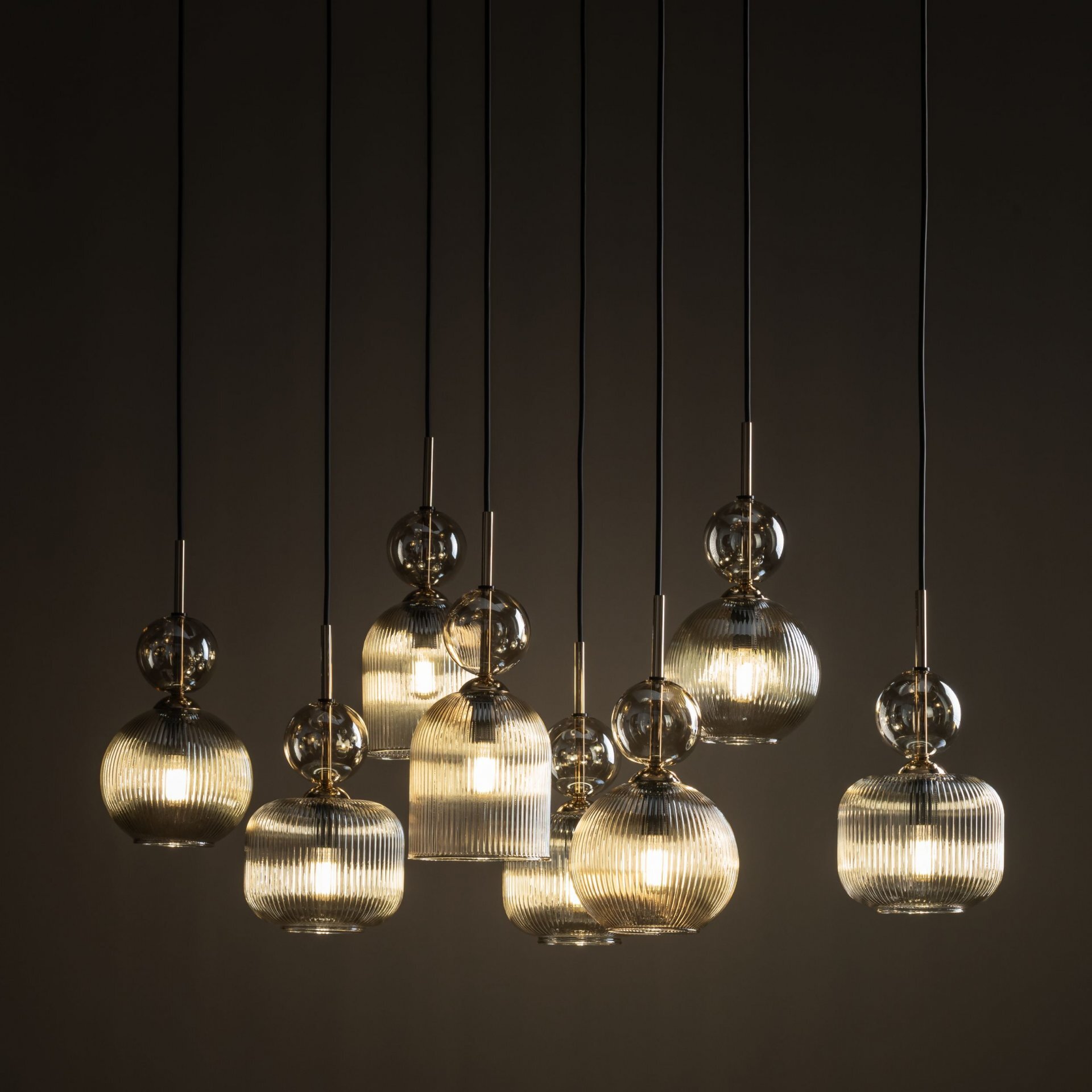 Na obrazie widoczna jest lampa wisząca z ośmioma szklanymi kloszami w kolorze koniakowym. Klosze mają żłobioną powierzchnię i są zawieszone na czarnych przewodach.