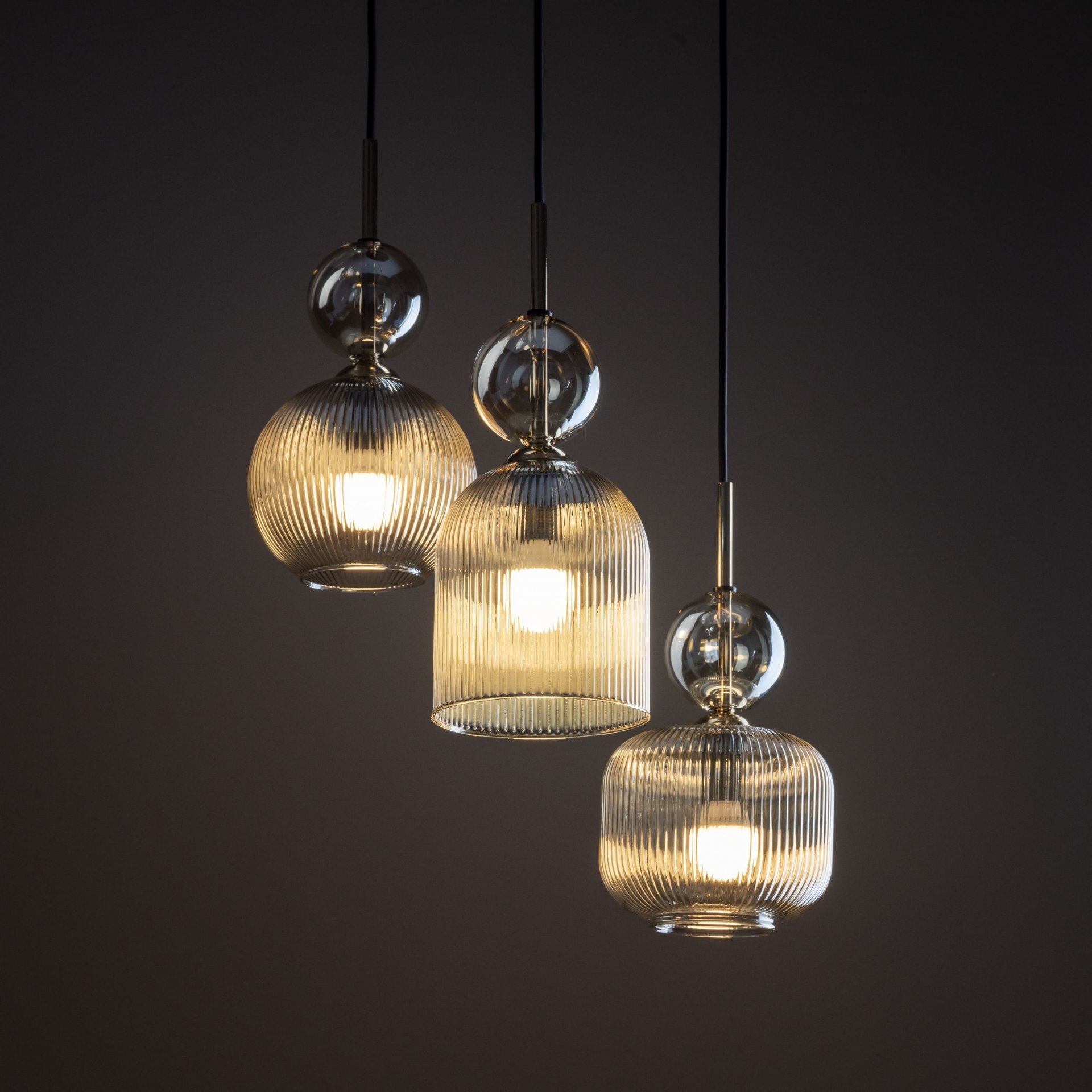 Trzy wiszące lampy z przezroczystymi, żebrowanymi kloszami, emitujące ciepłe światło. Każda lampa ma kulisty element dekoracyjny nad kloszem.