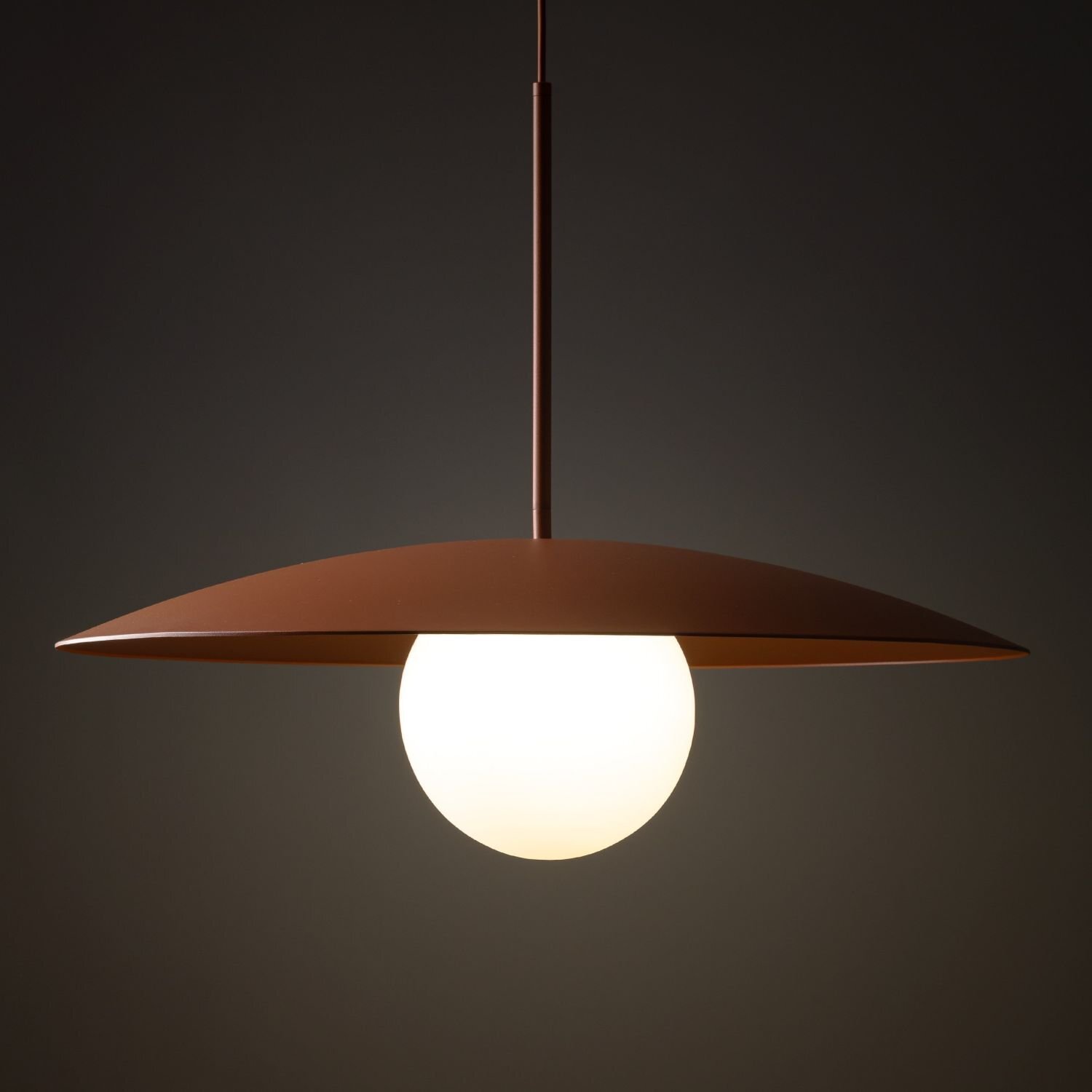 Lampa wisząca z okrągłym, białym kloszem i ceglastym, płaskim daszkiem. Lampa zawieszona jest na prostym przewodzie, a tło jest ciemne.