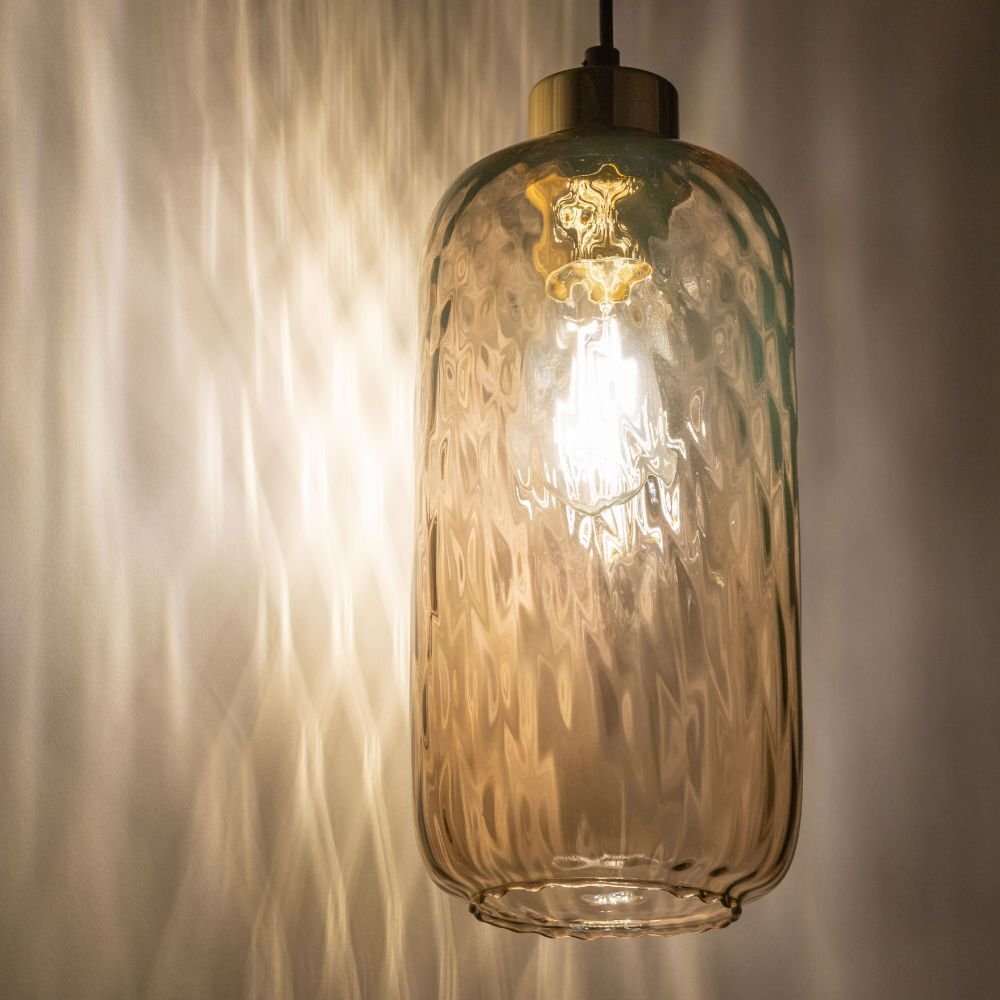 Lampa wisząca z bursztynowym szkłem, przez które prześwituje światło, tworząc wzory na ścianie. Klosz ma cylindryczny kształt z delikatnym wzorem i złotym wykończeniem u góry.