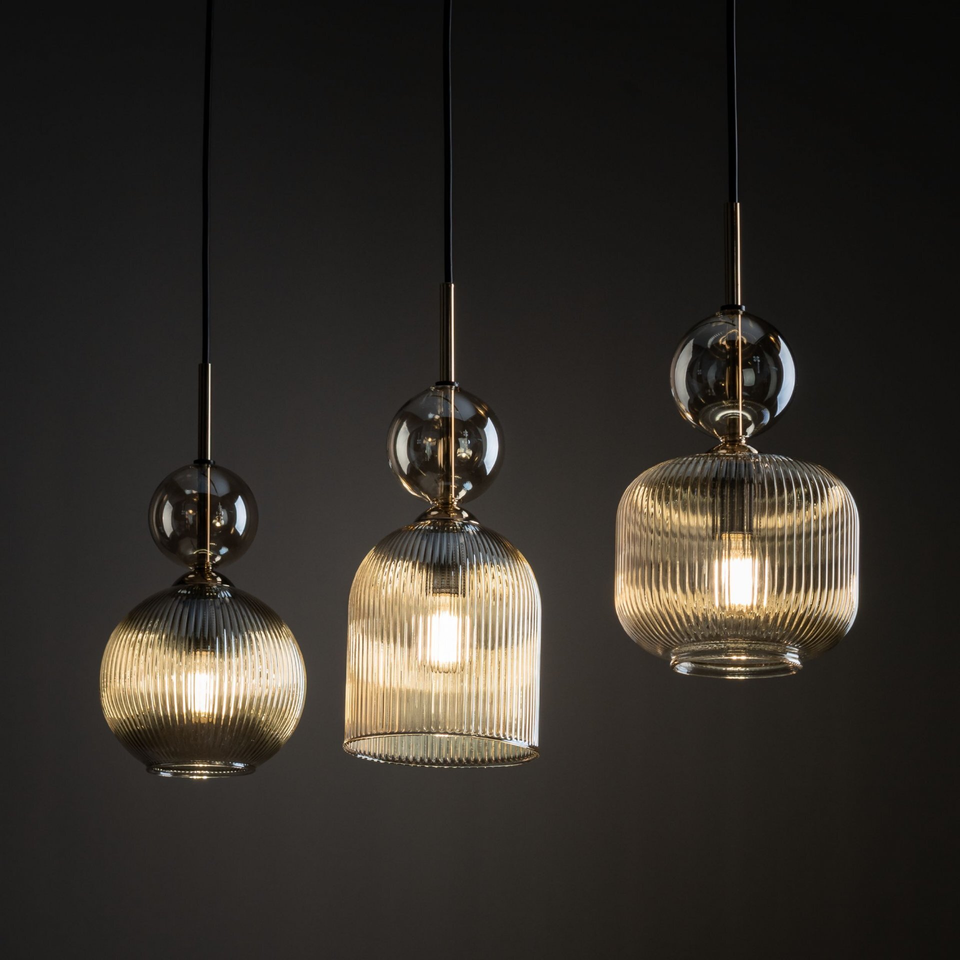 Trzy wiszące lampy z bursztynowymi, żebrowanymi kloszami i czarnymi przewodami, zawieszone na ciemnym tle. Każda lampa ma metalowy element łączący klosz z przewodem.