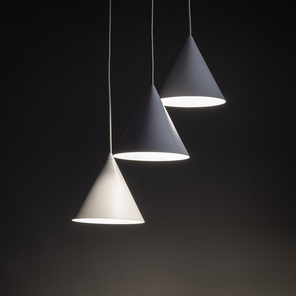Trzy białe, stożkowe lampy wiszące emitujące światło, zawieszone na cienkich przewodach na ciemnym tle. Lampy mają prosty, nowoczesny design.