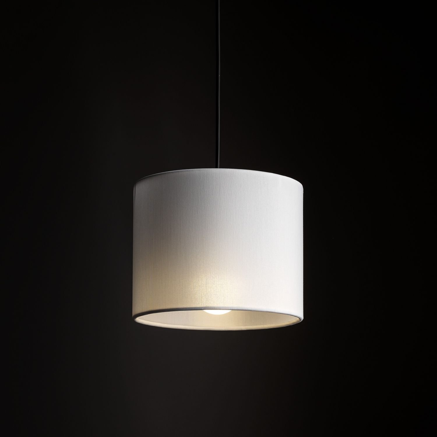 Na ciemnym tle wisi biała, cylindryczna lampa z widoczną żarówką wewnątrz. Lampa jest zawieszona na cienkim przewodzie.