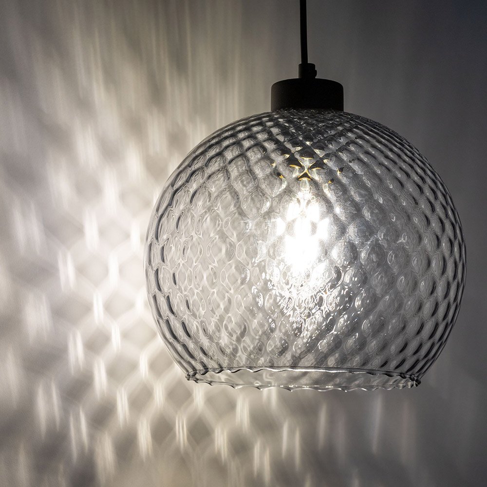 Szklana lampa wisząca z fakturą w kształcie heksagonów, emitująca światło tworzące wzory na ścianie. Lampa ma czarny uchwyt i jest zawieszona na kablu.