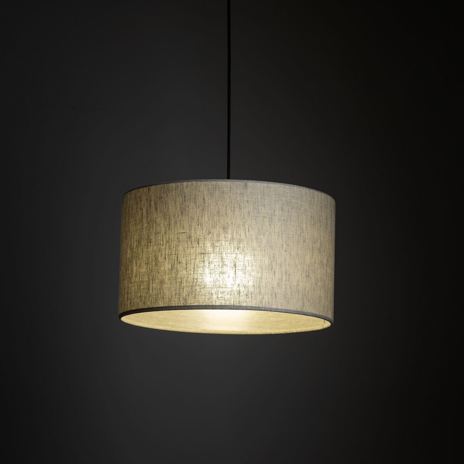 Na ciemnym tle wisi lampa z beżowym abażurem o cylindrycznym kształcie, emitująca ciepłe światło. Lampa jest zawieszona na cienkim czarnym przewodzie.