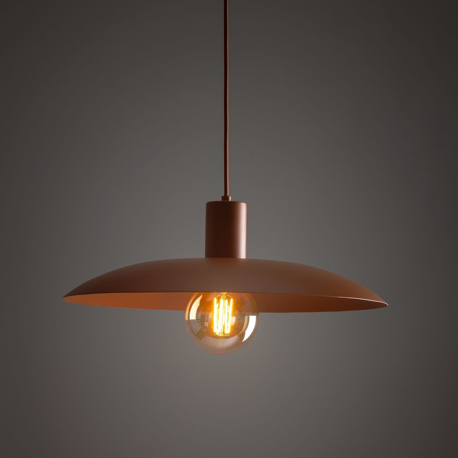Lampa wisząca w kolorze ceglasto-pomarańczowym z metalowym kloszem i widoczną żarówką wewnątrz. Lampa zawieszona jest na cienkim przewodzie, a tło jest ciemne.
