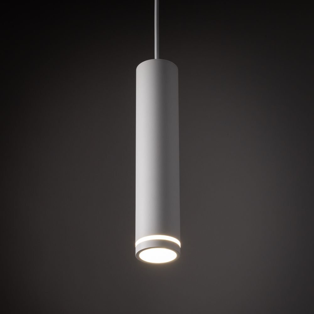 Biała, cylindryczna lampa wisząca z metalu, emitująca światło z dolnej części. Lampa zawieszona jest na cienkim przewodzie, na ciemnym tle.