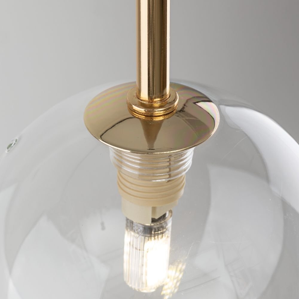 Zbliżenie na przezroczysty klosz lampy z widoczną żarówką i złotym elementem mocującym. Lampa ma elegancki, nowoczesny design.
