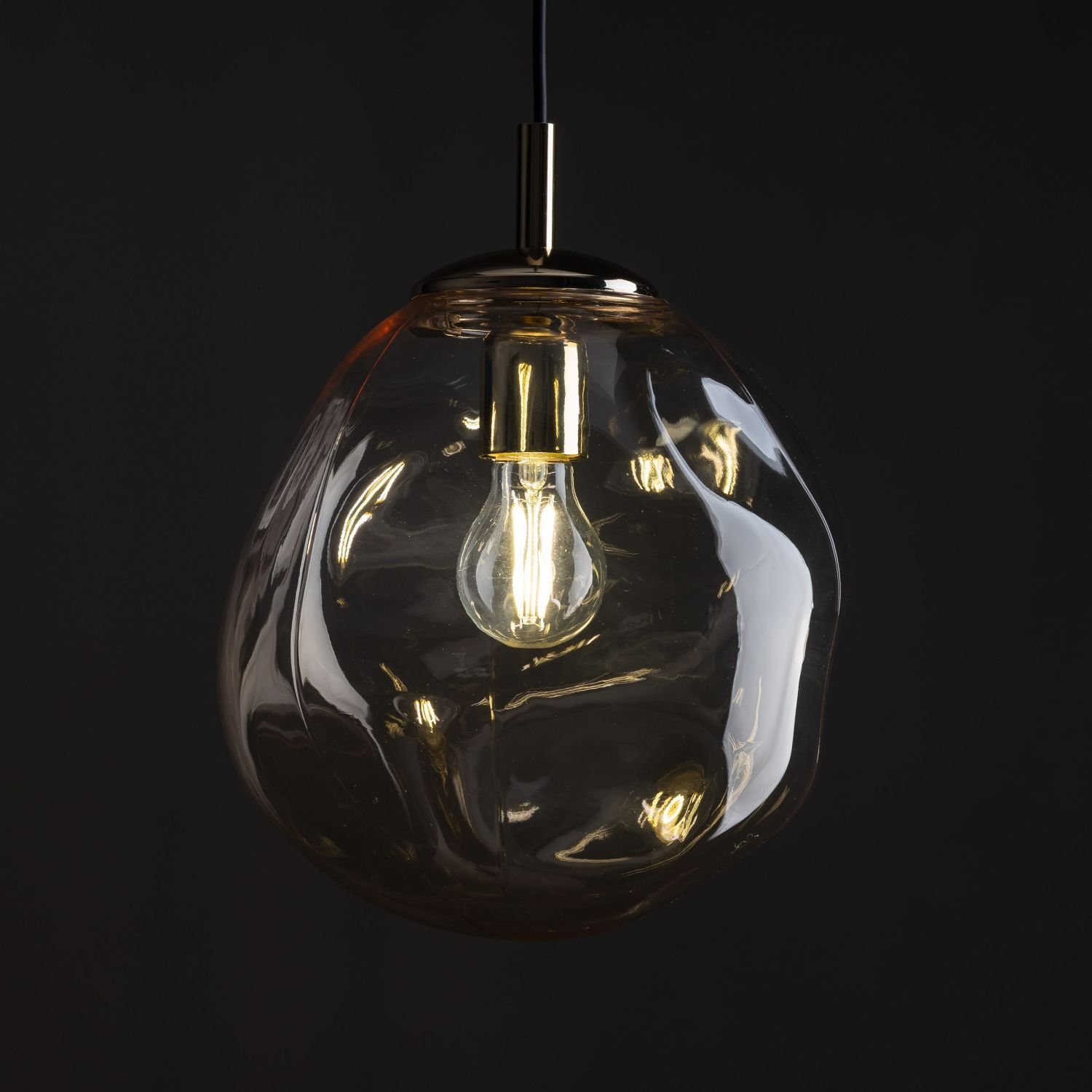 Lampa wisząca z bursztynowym kloszem o nieregularnym kształcie, wewnątrz której znajduje się żarówka. Elementy metalowe są w kolorze czarno-złotym.