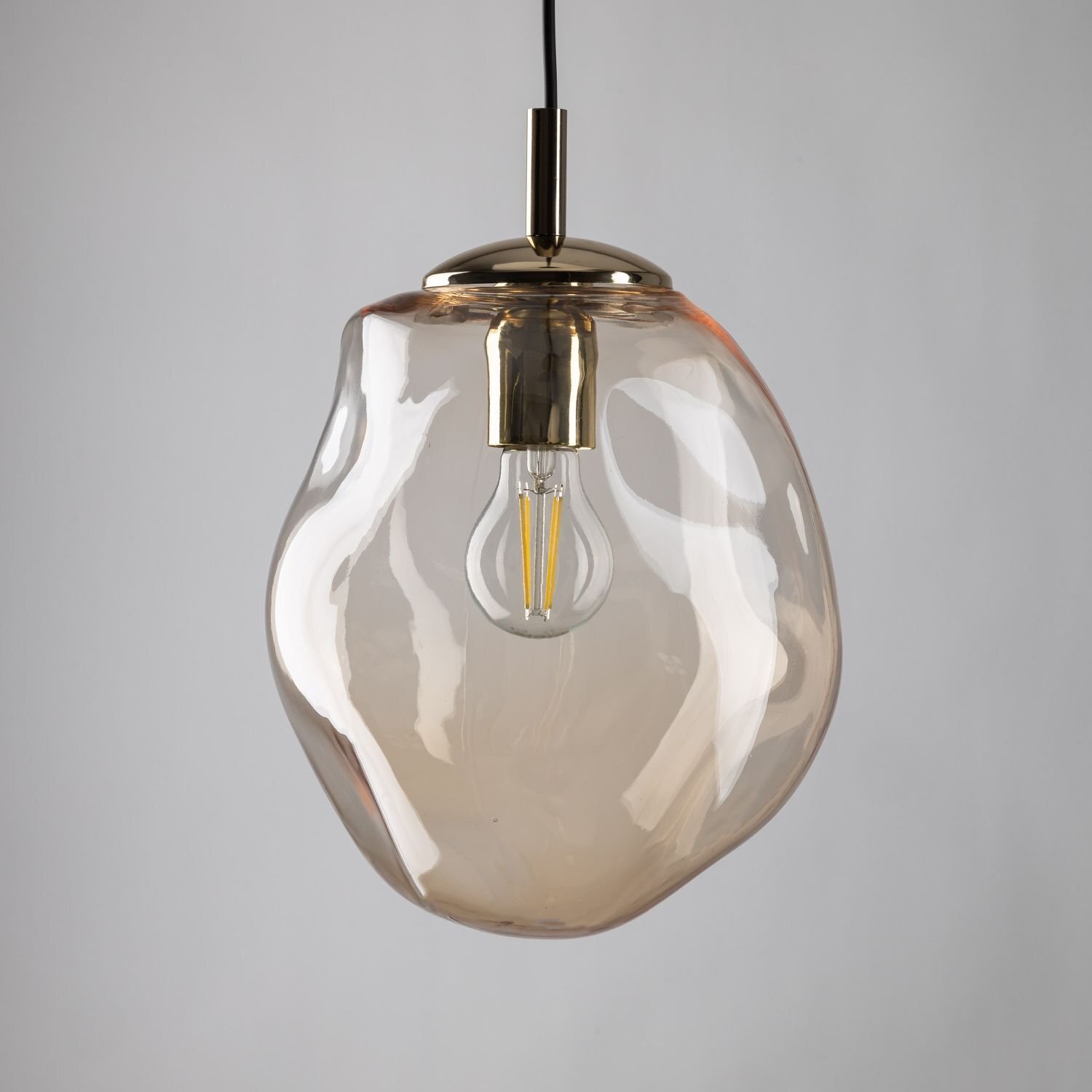 Lampa wisząca z bursztynowym kloszem o nieregularnym kształcie, z widoczną żarówką wewnątrz. Elementy metalowe są w kolorze złotym, a przewód jest czarny.