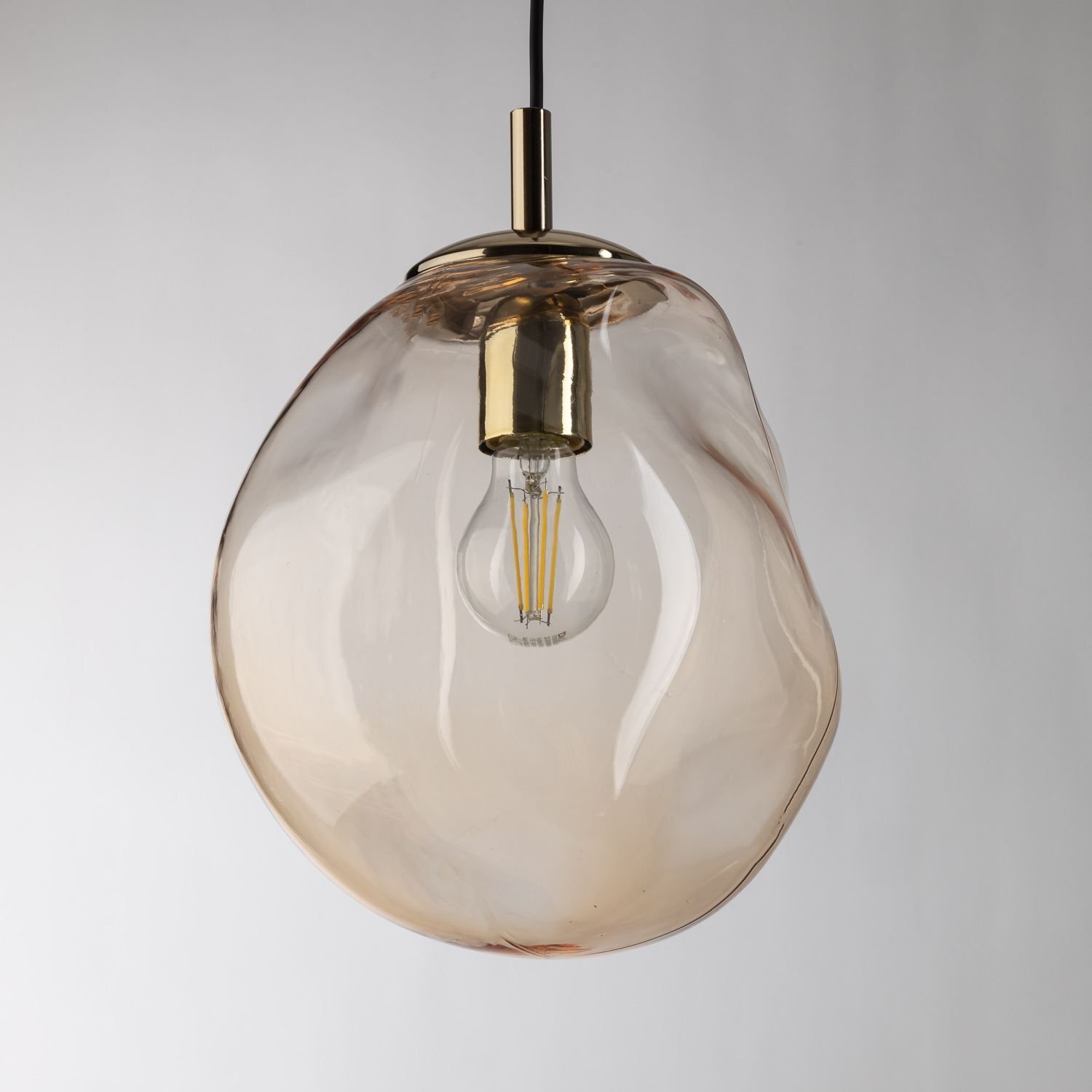 Lampa wisząca z bursztynowym kloszem o nieregularnym kształcie, z widoczną żarówką wewnątrz. Elementy mocujące są w kolorze złotym.