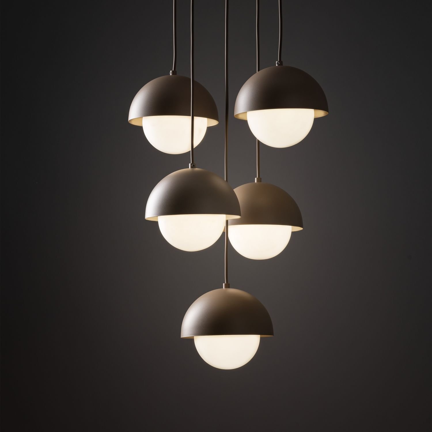 Na obrazie widoczna jest lampa wisząca z pięcioma kloszami w kształcie półkul, w kolorze beżowym. Klosze emitują ciepłe, białe światło, a całość zawieszona jest na ciemnym tle.