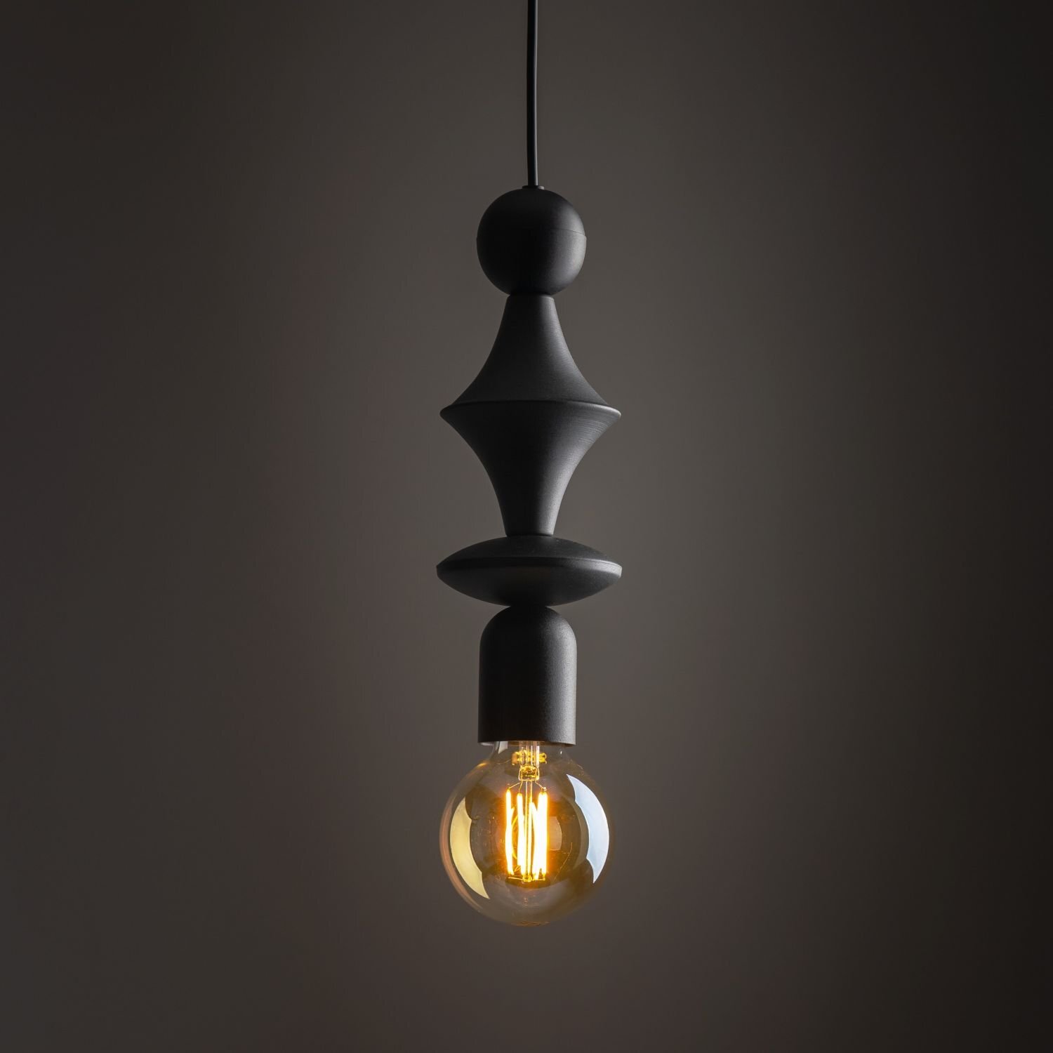 Czarna, metalowa lampa wisząca z ozdobnym trzonkiem i widoczną żarówką emitującą ciepłe światło. Lampa ma nowoczesny design i jest zawieszona na ciemnym tle.