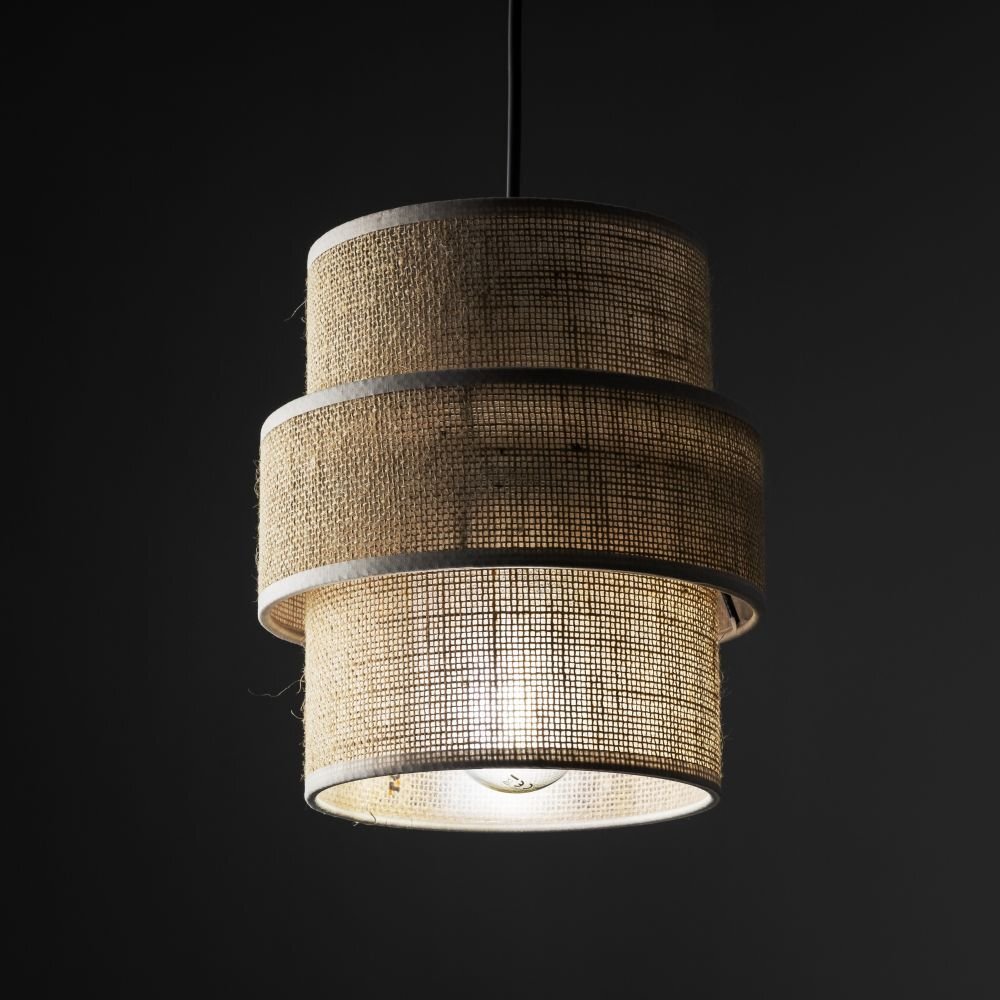 Lampa wisząca z trzema cylindrycznymi abażurami wykonanymi z beżowej juty, z widoczną żarówką wewnątrz. Elementy konstrukcyjne są czarne, a tło jest ciemne.