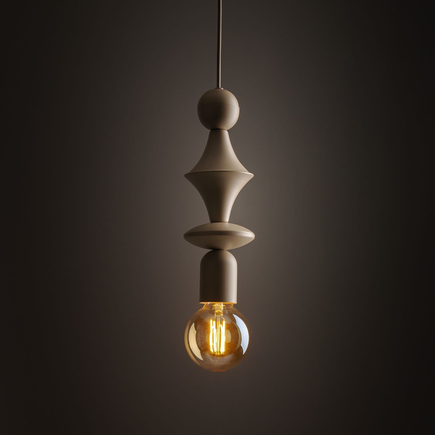 Beżowa lampa wisząca o nowoczesnym designie z widoczną żarówką emitującą ciepłe światło. Lampa ma kształt tuby z dekoracyjnymi elementami i jest zawieszona na cienkim przewodzie.
