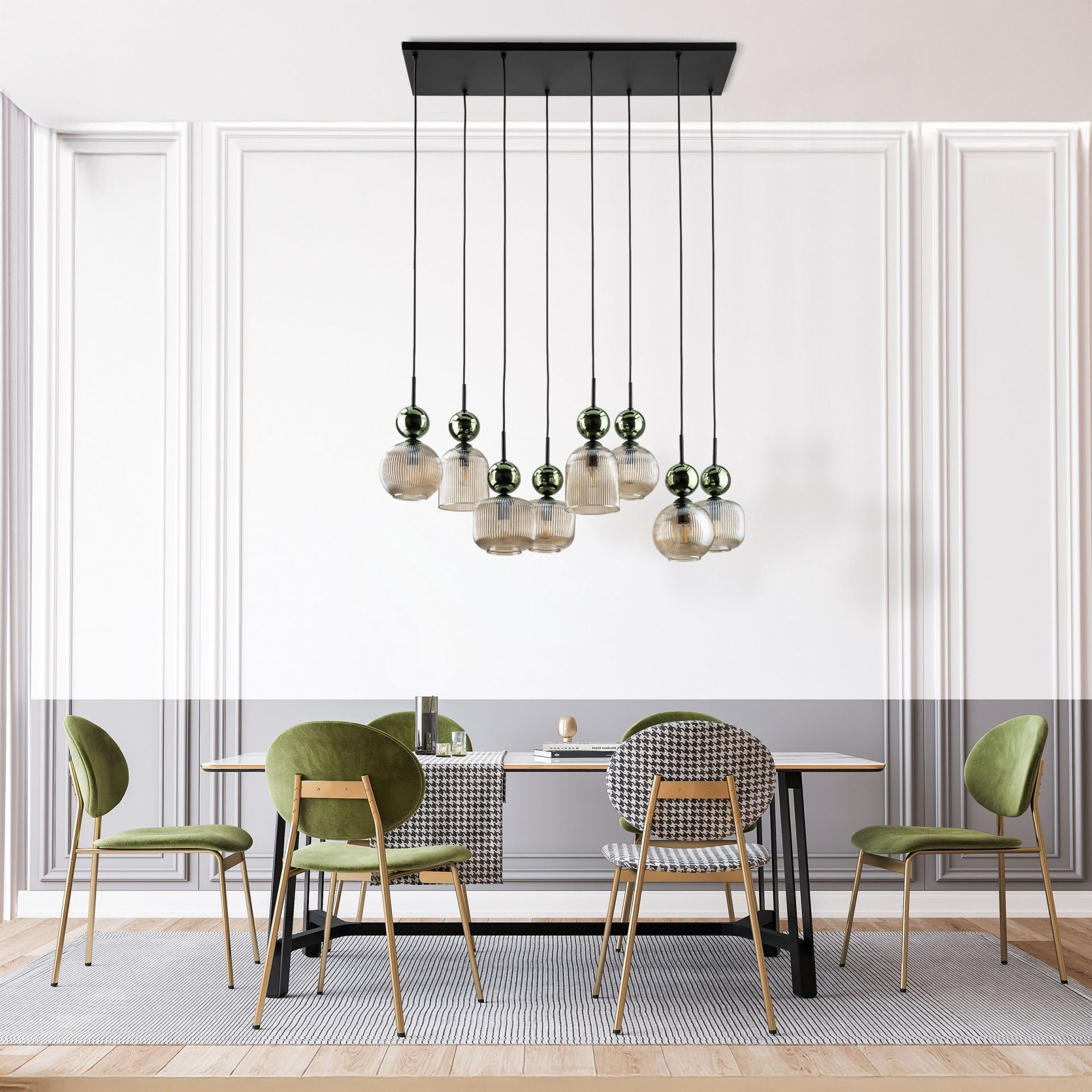 Wnętrze z elegancką lampą wiszącą składającą się z ośmiu szklanych kloszy w kolorach zielonym i koniakowym, zawieszonych na czarnej konstrukcji. Pod lampą znajduje się stół otoczony zielonymi krzesłami na drewnianej podłodze.