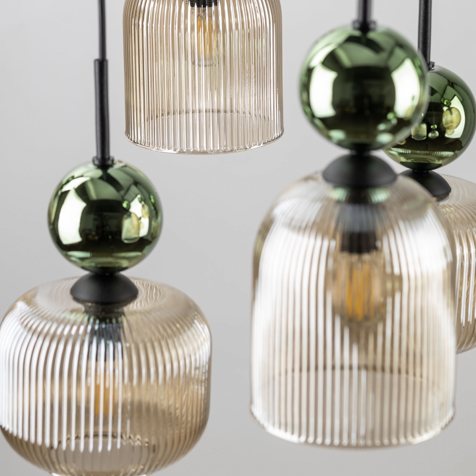 Zdjęcie przedstawia zbliżenie na kilka szklanych kloszy lamp wiszących o żebrowanej strukturze, w kolorze bursztynowym i zielonym. Klosze są zawieszone na czarnych konstrukcjach.