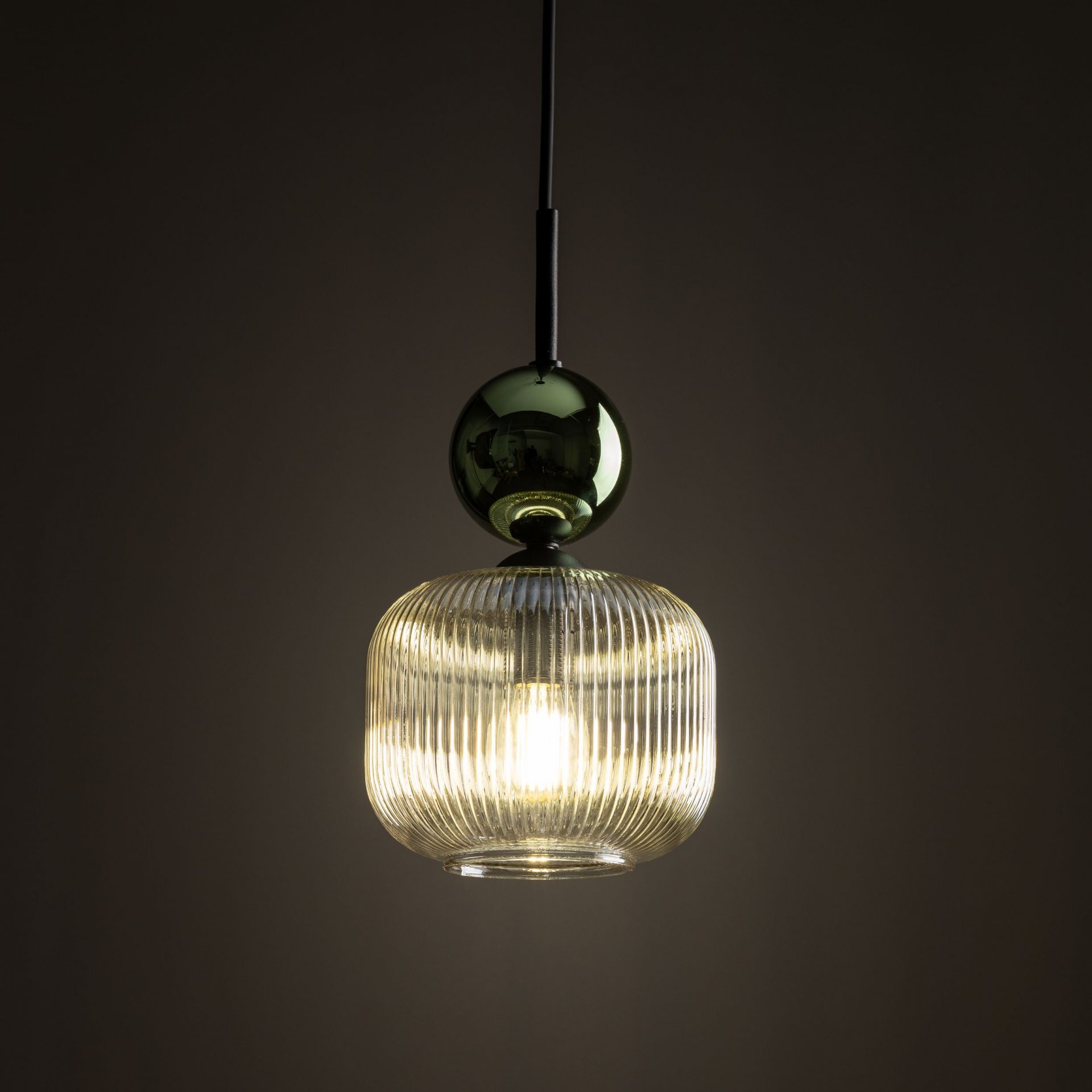 Lampa wisząca z żarówką LED, posiada szklany klosz o żłobionej powierzchni i ciemnozieloną kulę nad nim. Światło emituje ciepły blask, a całość zawieszona jest na czarnym przewodzie.