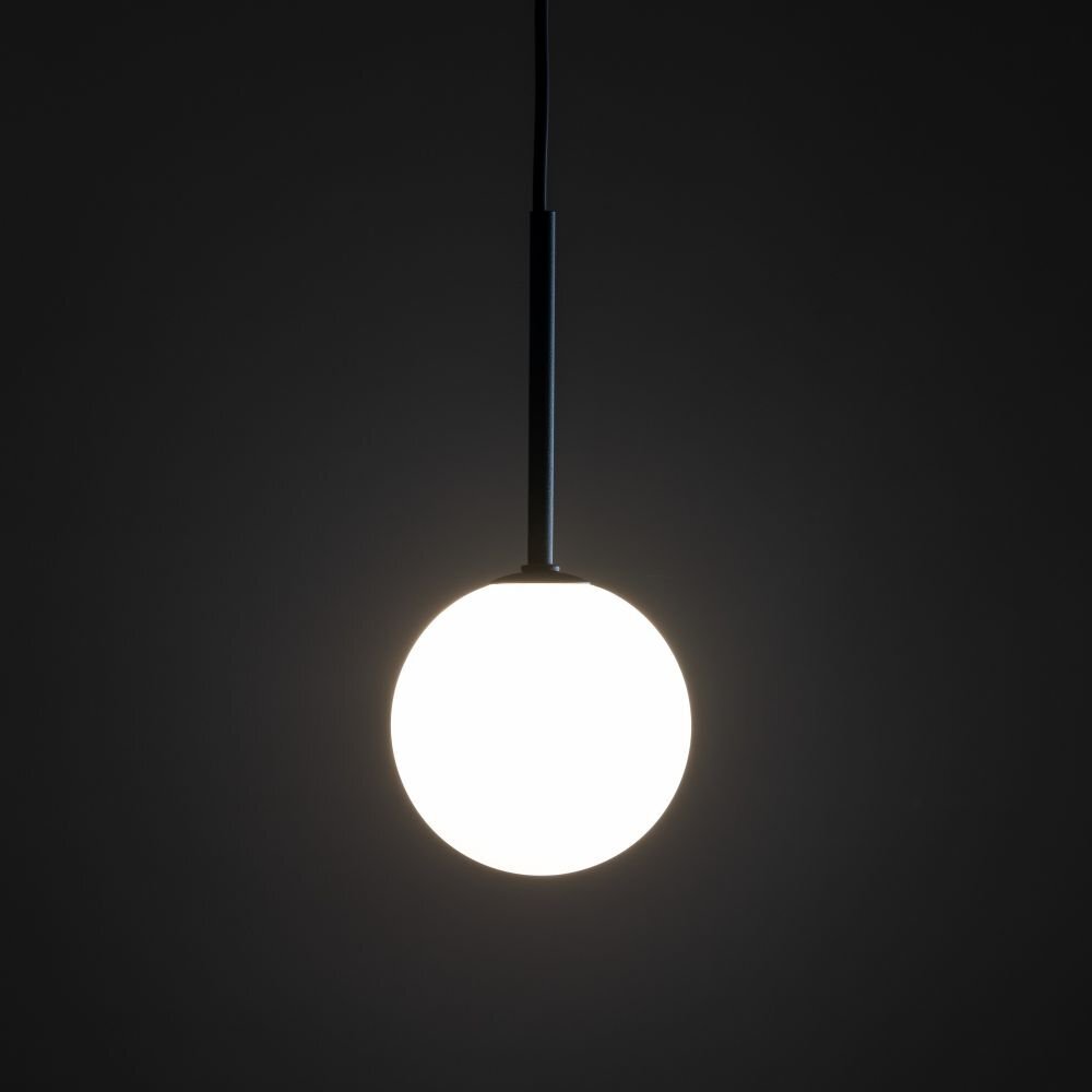 Na ciemnym tle wisi lampa z okrągłym, białym kloszem i czarnym uchwytem. Światło lampy jest włączone, tworząc jasną poświatę wokół klosza.