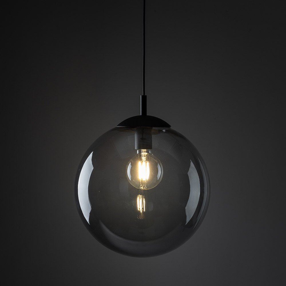 Na ciemnym tle wisi lampa z przezroczystym, kulistym kloszem, wewnątrz którego znajduje się żarówka emitująca ciepłe światło. Lampa ma prosty, nowoczesny design z grafitowym wykończeniem.