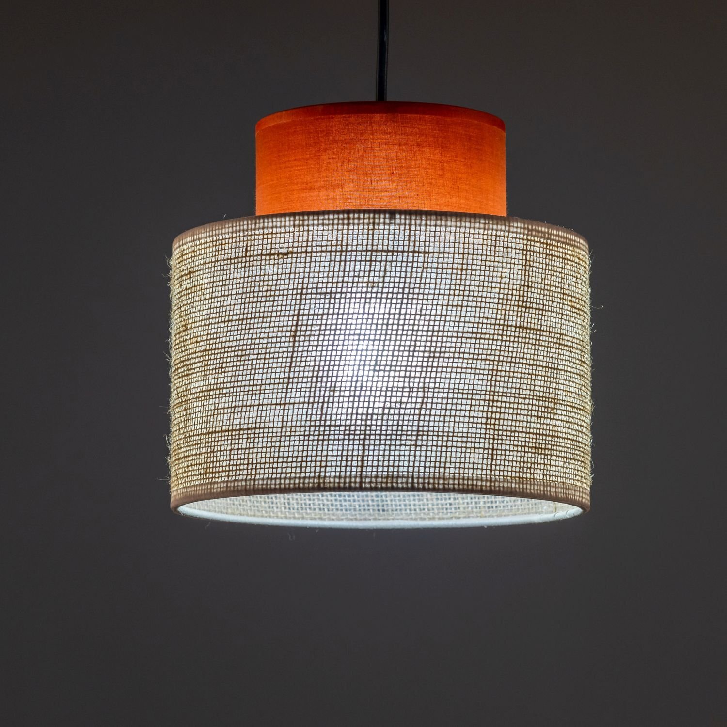 Lampa wisząca z cylindrycznym kloszem wykonanym z naturalnej juty, z wewnętrznym elementem w kolorze pomarańczowym. Światło przenika przez teksturę materiału, tworząc ciepły efekt świetlny.