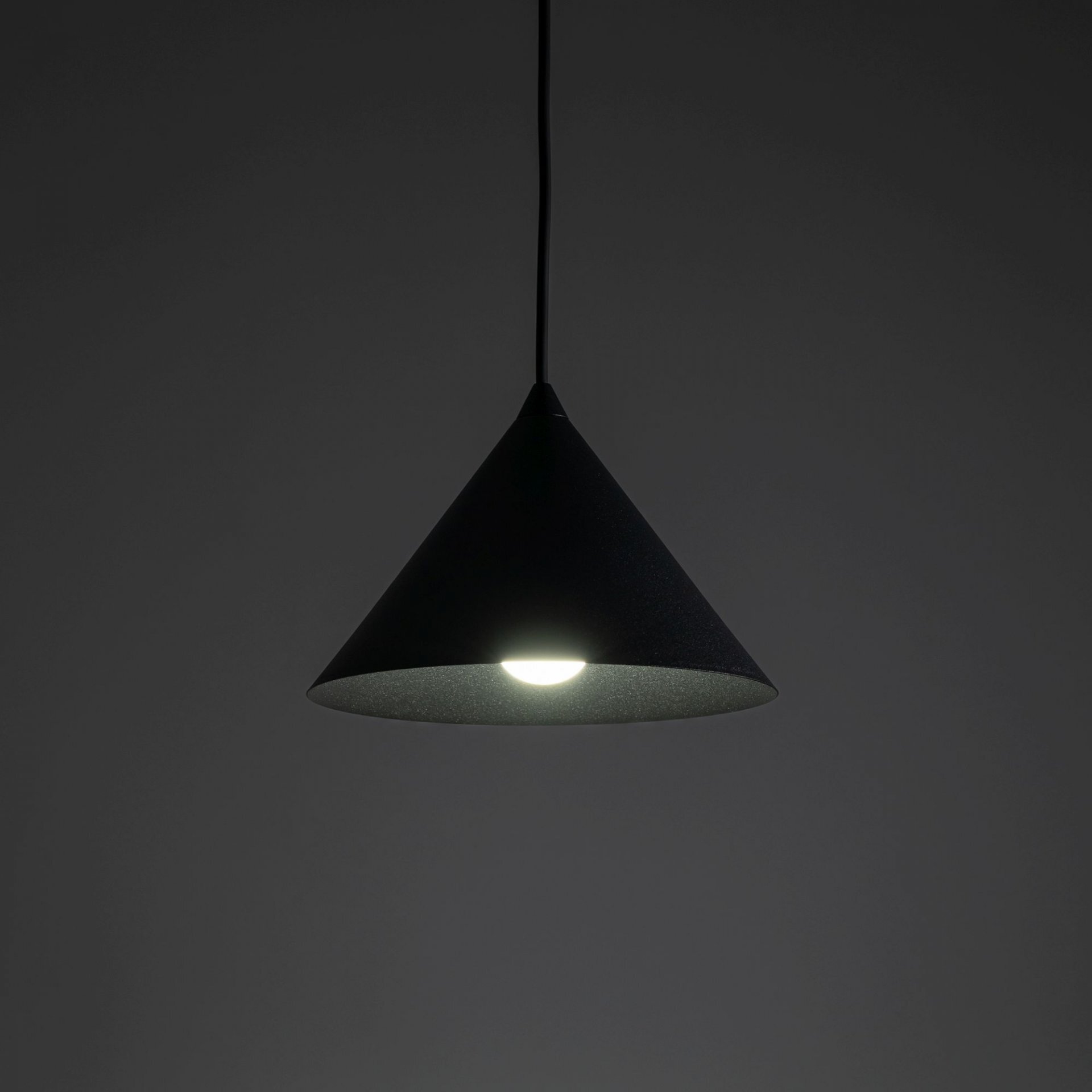 Czarna, stożkowa lampa wisząca z metalowym kloszem, emitująca jasne światło. Lampa zawieszona jest na cienkim przewodzie, na ciemnym tle.