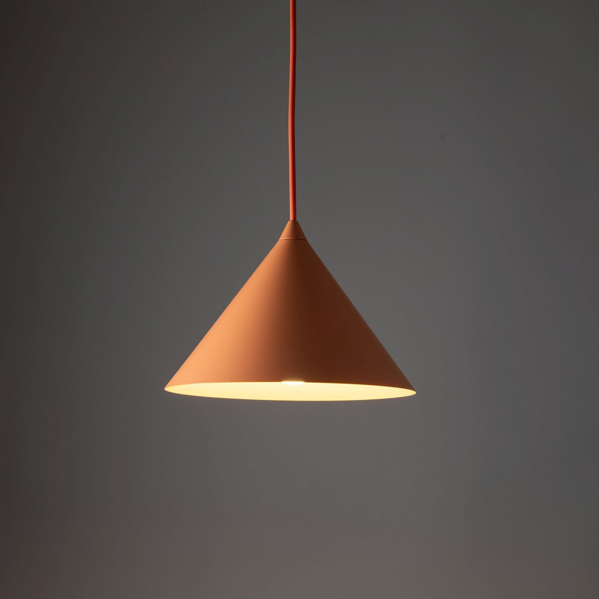 Pomarańczowa lampa wisząca o stożkowym kształcie, z widocznym przewodem w tym samym kolorze. Lampa emituje ciepłe światło na ciemnym tle.
