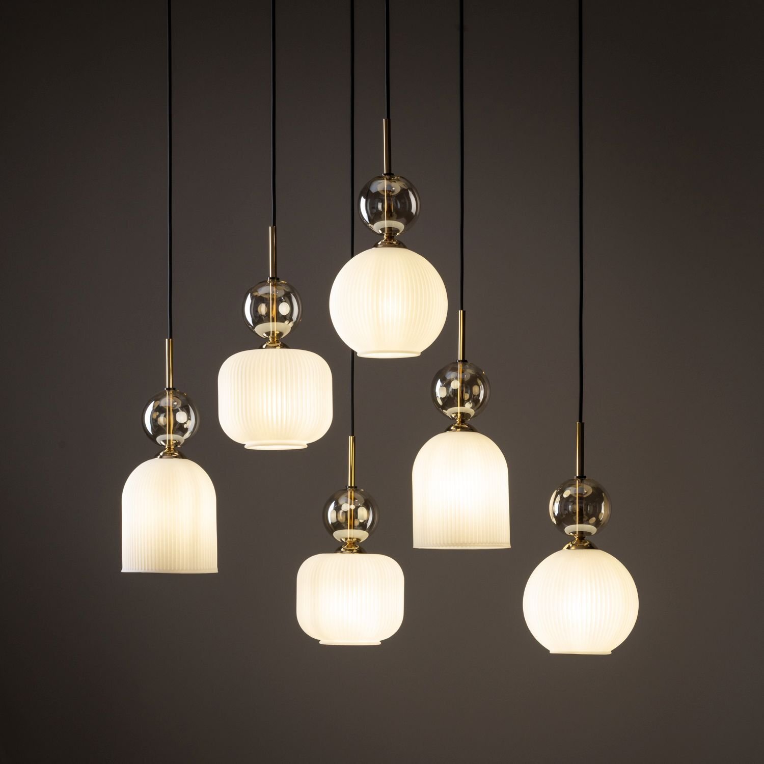 Na obrazie widoczna jest lampa wisząca z sześcioma kloszami w kolorze białym, zawieszonymi na czarnych przewodach. Klosze mają różne kształty i są ozdobione złotymi elementami oraz przezroczystymi kulami.